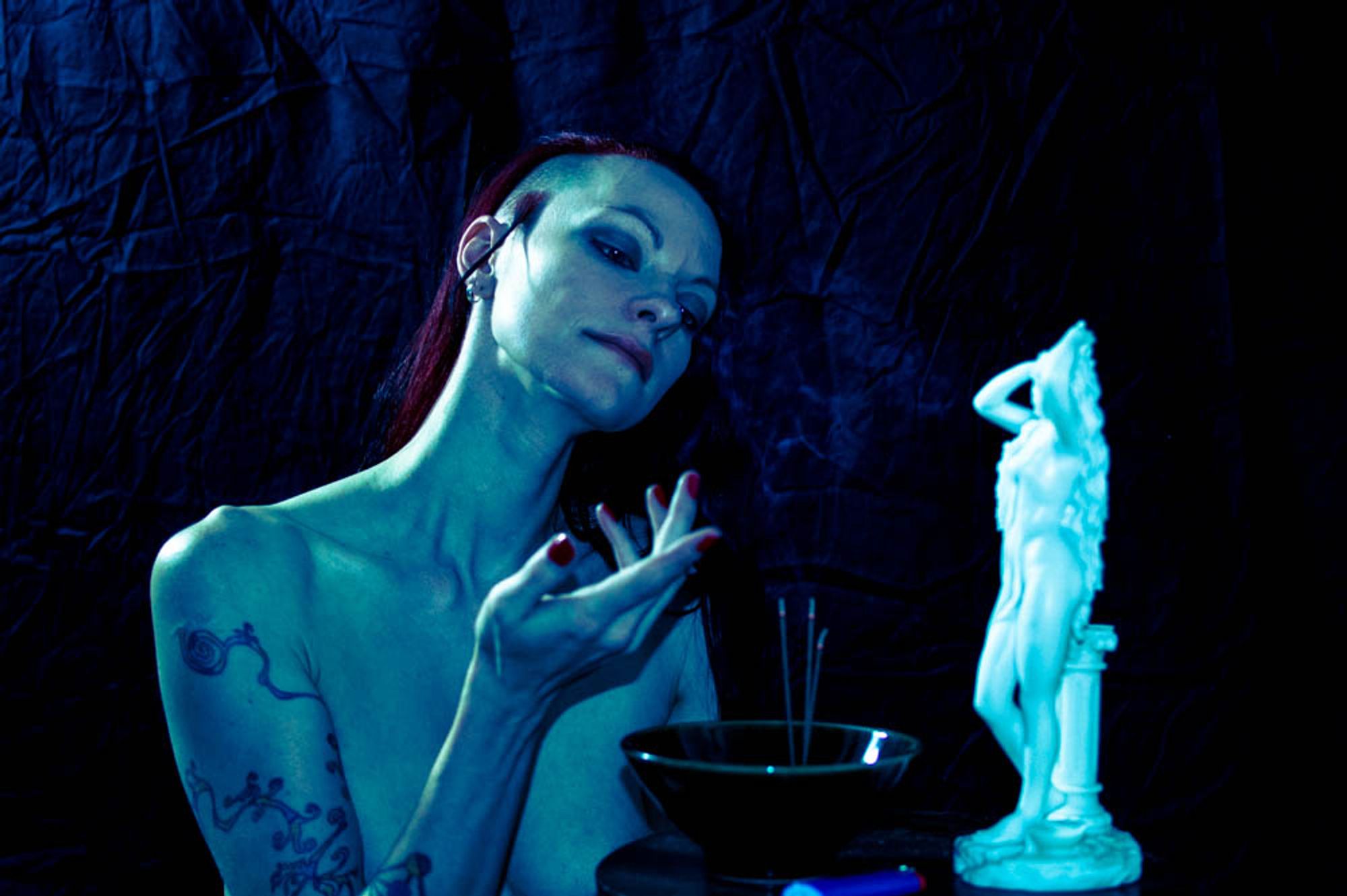 a scrawny white woman kneels behind an altar. before her is a bowl of incense sticks and an idol of venus