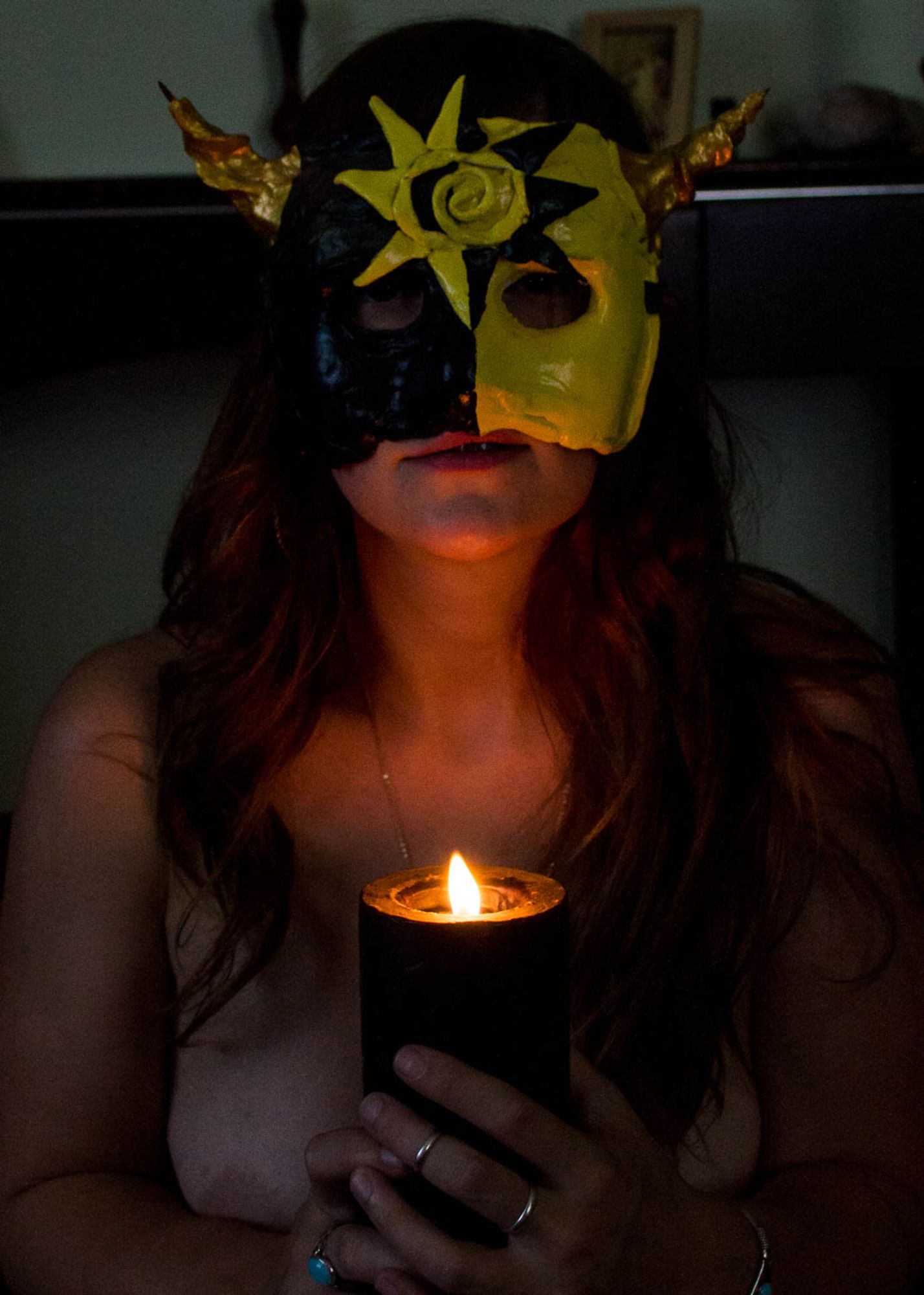 photo of a young white woman with long dark hair, wearing a black and yellow solar mask and staring at the viewer. Her face is illuminated by a black candle