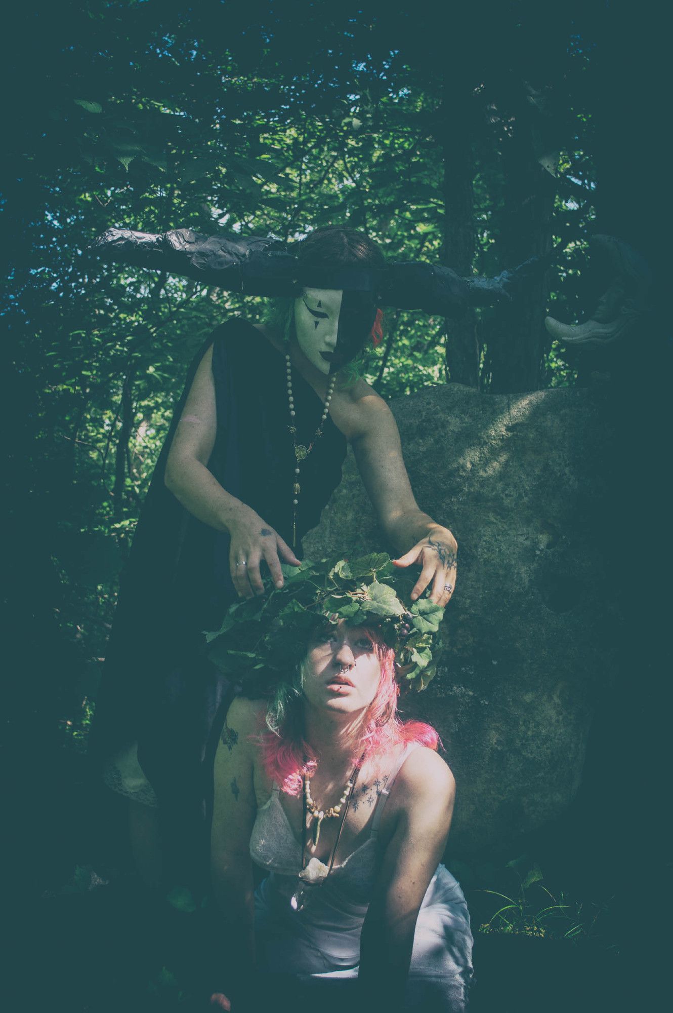 matte color photo. a shadowy horned and masked figure in a black chiton looms behind a kneeling young woman with pink hair and a crown of grape vines. The figure is reaching down to the woman's head even as the woman's eyes turn heavenward.