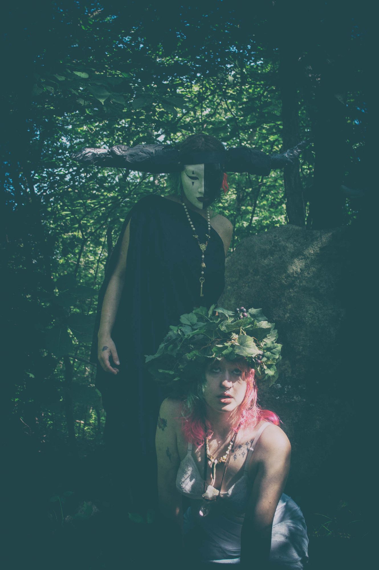 matte color photo. a shadowy horned and masked figure in a black chiton looms behind a kneeling young woman with pink hair and a crown of grape vines