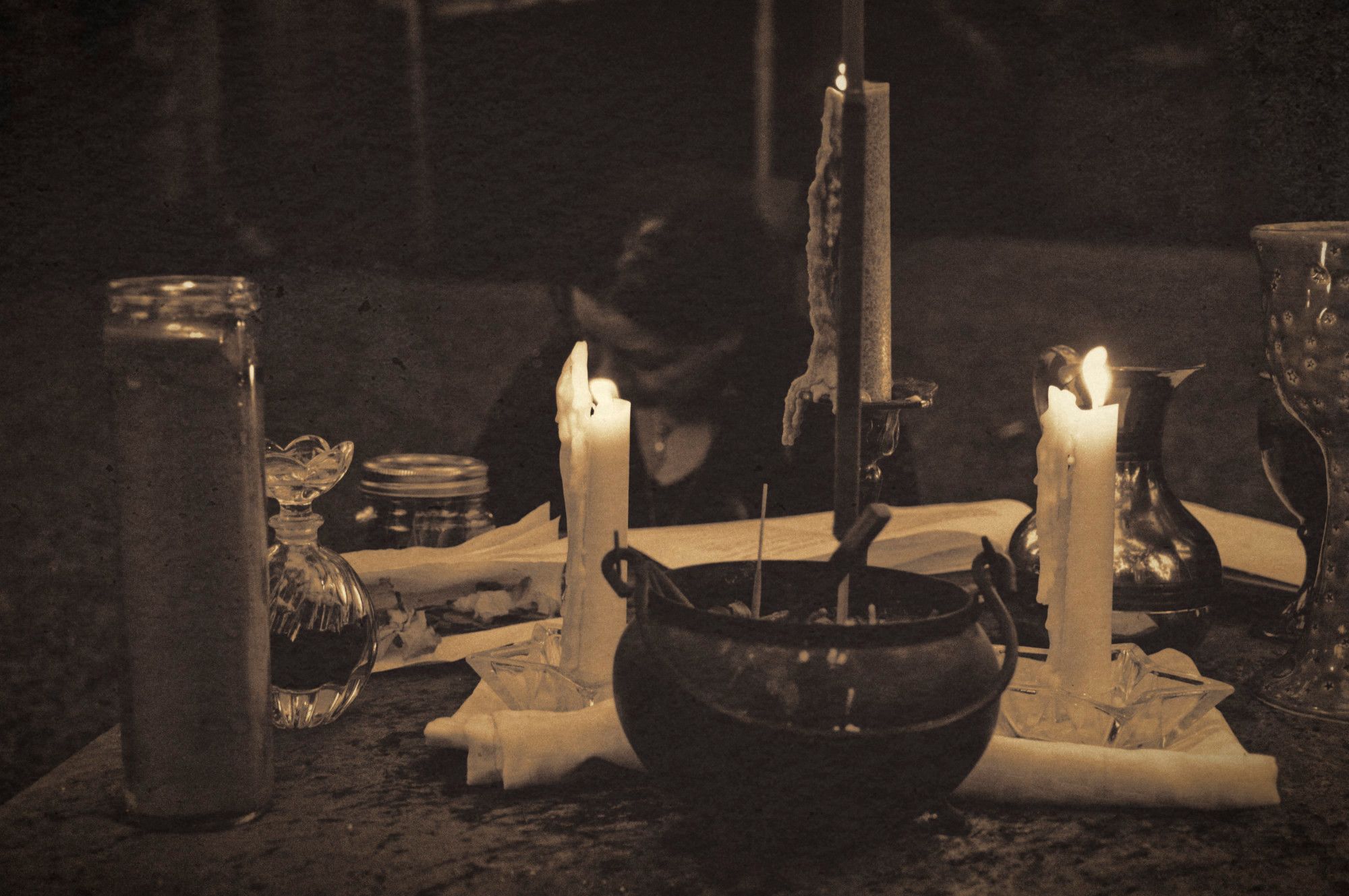 a sepia tone photo of a candlelit altar. in the background, just out of focus, a woman kneels as she performs her rites