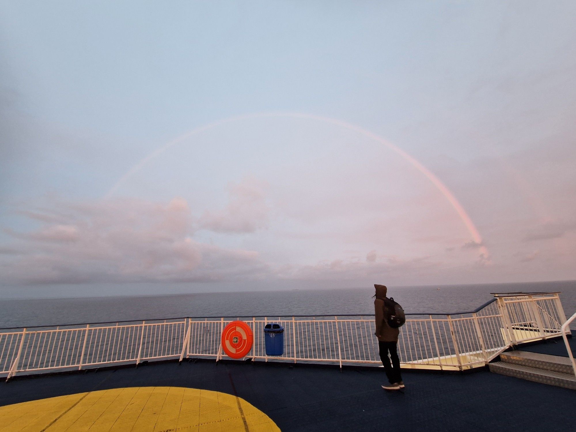 A view from the top deck of a cruise ship, a rainbow is visible through the slightest overcast sky