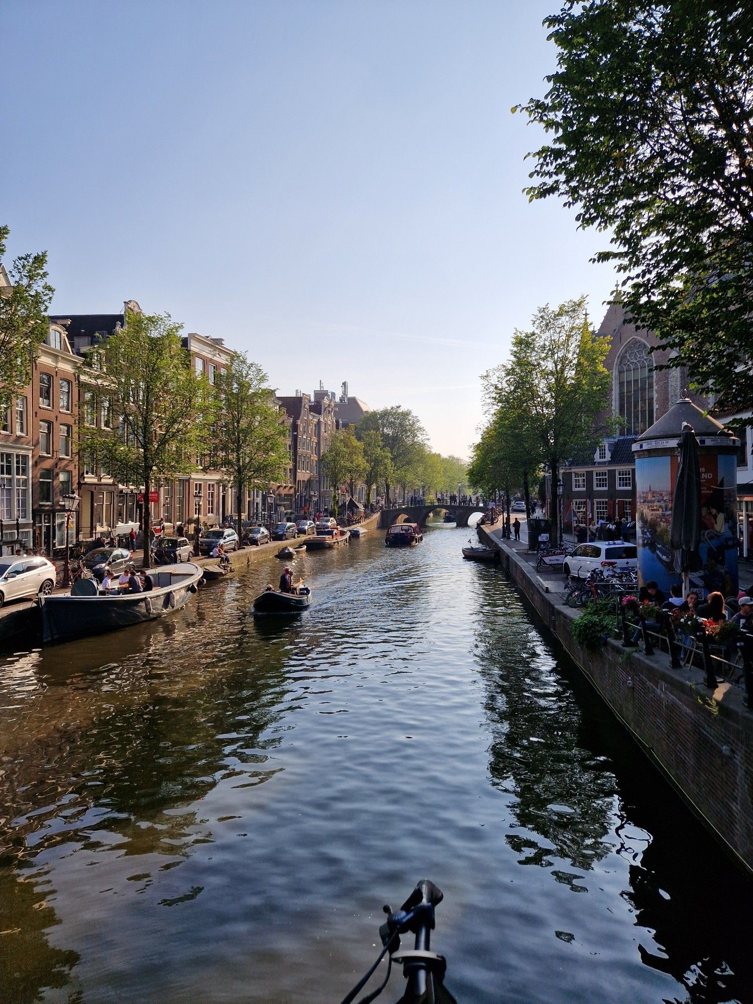 Photo of a canal in Amsterdam. There's a boat going towards the bridge, either side of the canal up have houses and cards parked dangerously close to the water.