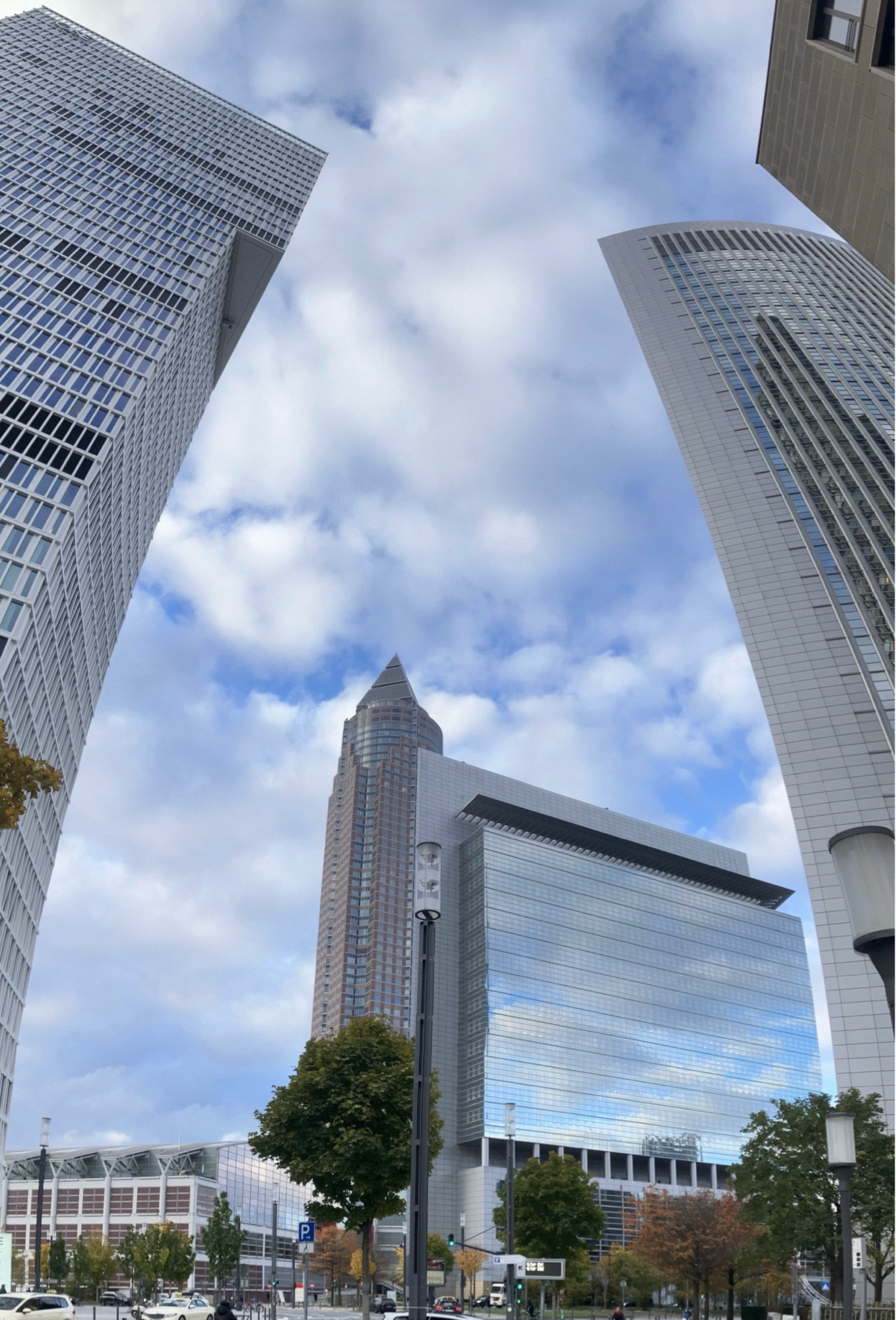 Frankfurt- Pano-Aufnahme 2er Hochäuser, die sich durch die Verzerrung der Kamerabewegung zueinanderneigen, dahinter der Messeturm, Bäume, ein weiteres Hochhaus, dessen Fassade den Schäfchenwolkenhimmel spiegelt.