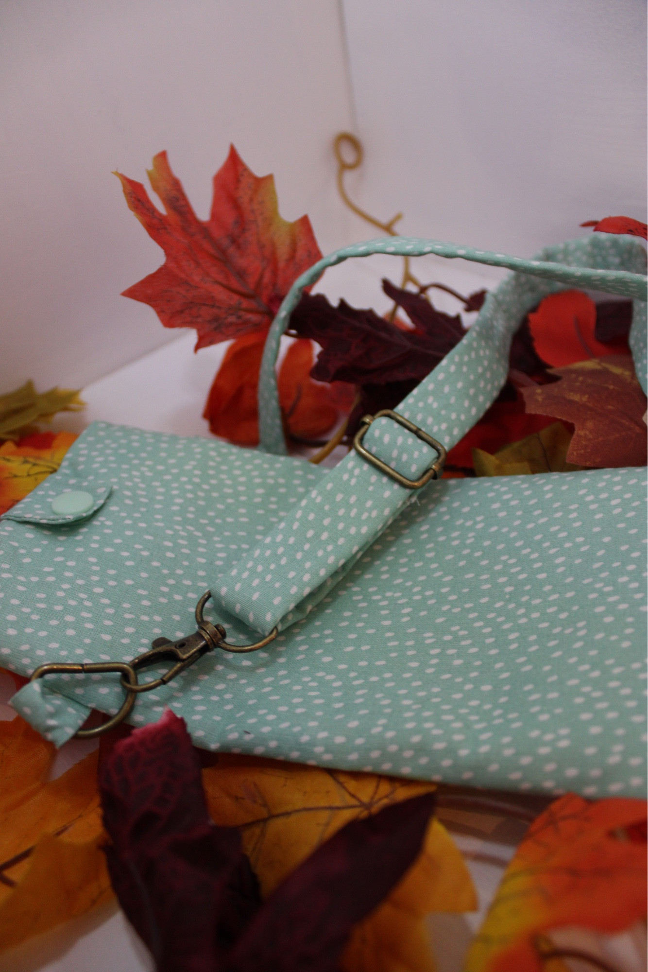 Pochette à téléphone, cousue dans un tissu bleu turquoise à pois blanc. 
La pochette possède une lanière réalisée dans le même tissu et se referme avec un bouton pression bleu turquoise 
On voit notamment la lanière réglable de plus près