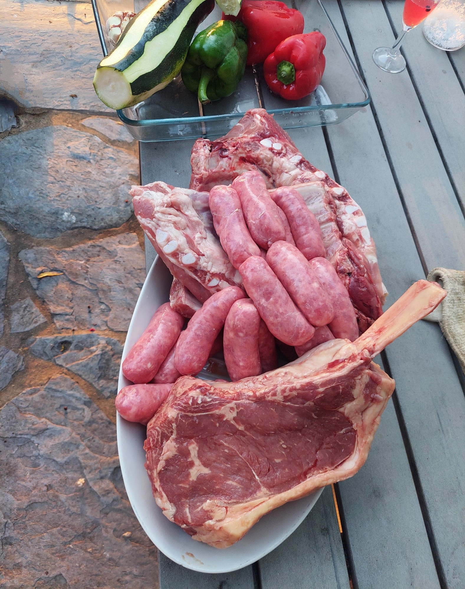 Auf einem Holztisch im Fteien steht eine Platte mit rohwm Fleisch und Würstchen und eine weitere mit verschiedenem Gemüse.