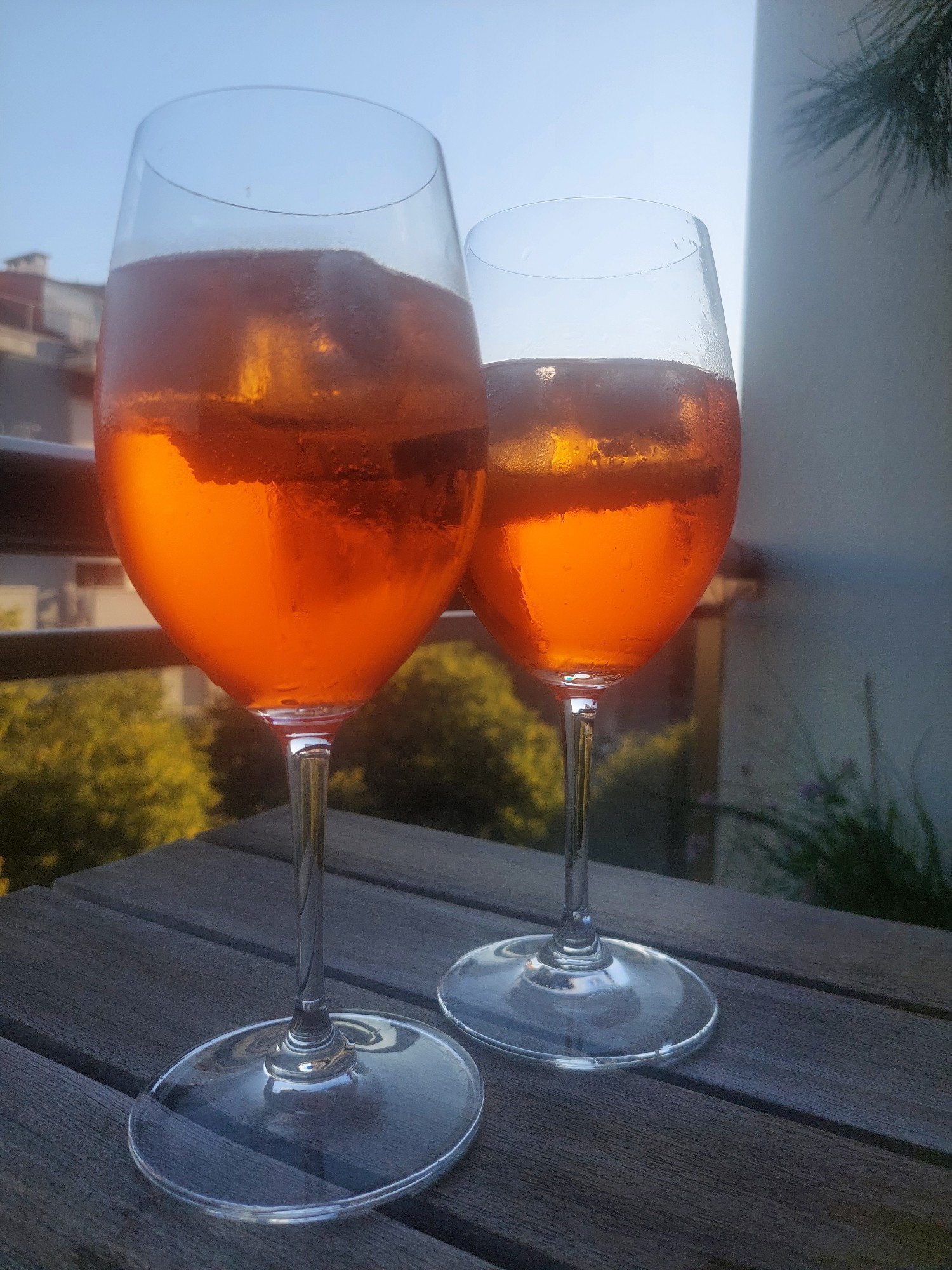 Zwei Gläser mit einem orangefarbenen Getränk stehen auf einem Holztisch auf einem Balkon, im Hintergrund Bäume und eine Hauswand