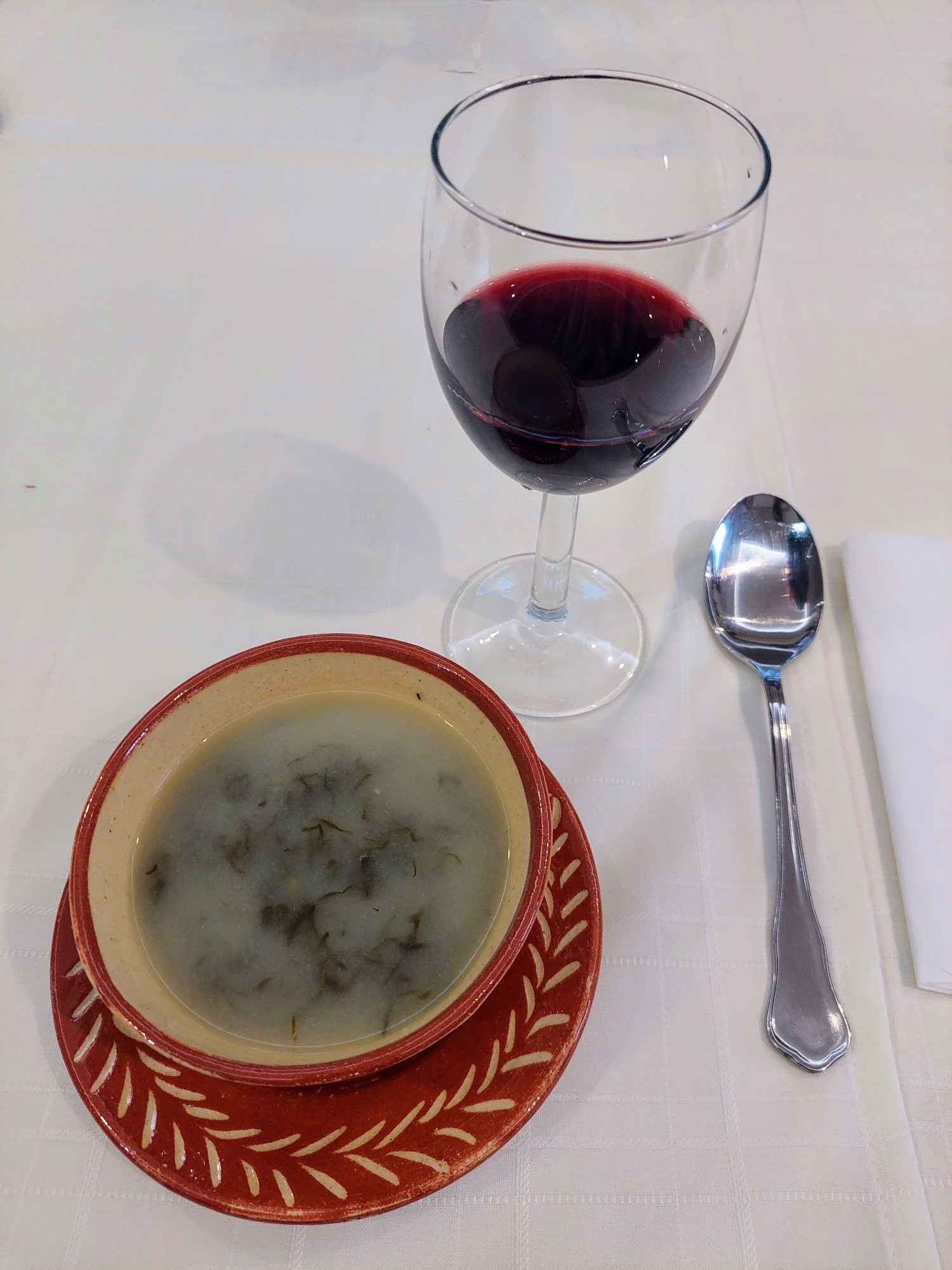 Auf einem mit Tischdecke bedeckten Tisch stehen bzw. liegen die folgenden Hilfsmittel: Serviette, Löffel, Weinglas (mit Rotwein), Suppentasse (mit Suppe)