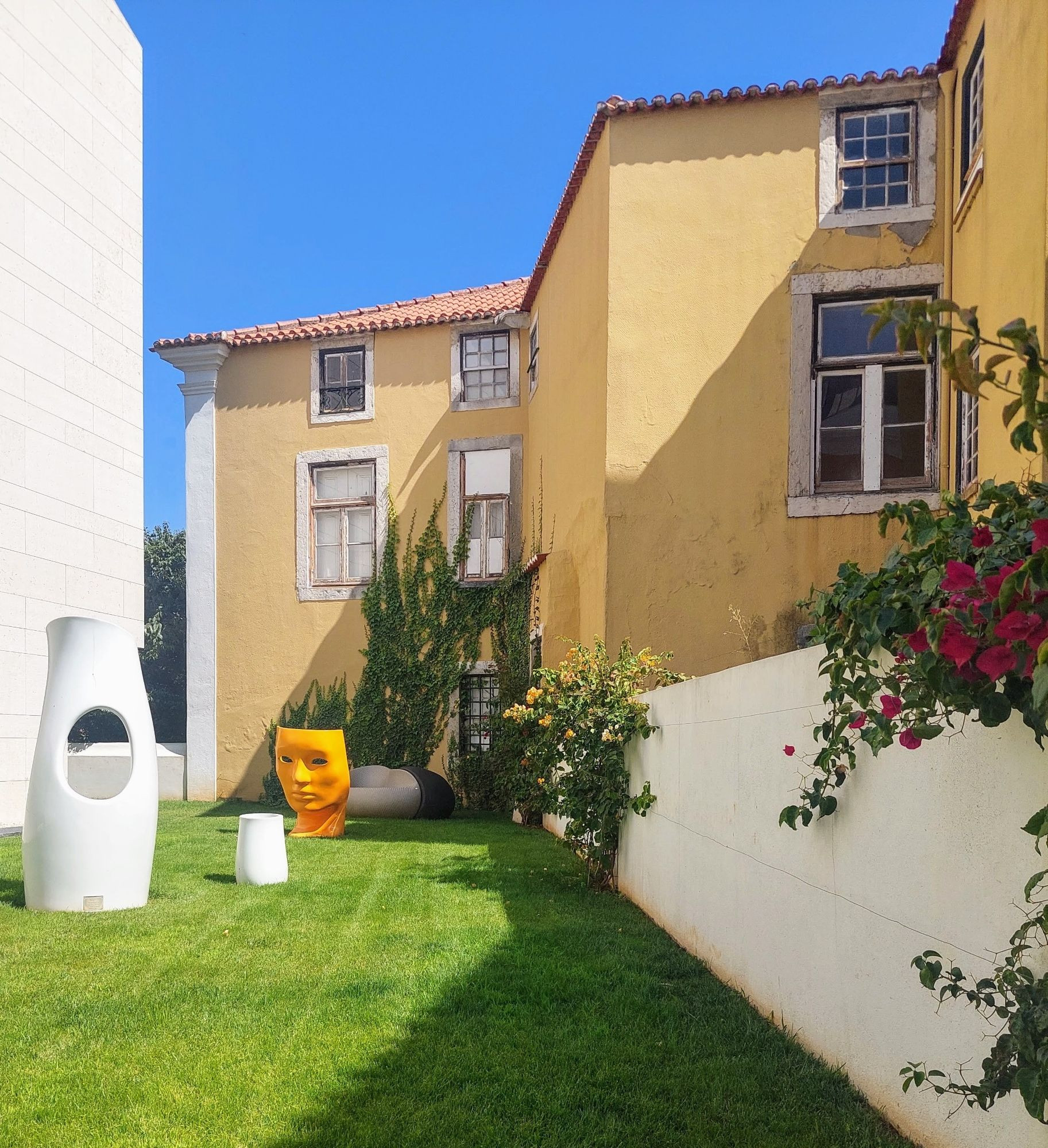 Gartenbereich, der von Gebäuden mit ockerfarbenen Wänden und weißen Fensterrahmen umgeben ist, darüber blauer Himmel. Im Vordergrund befinden sich eine große und eine kleine weiße Skulptur. Daneben liegt eine kleinere Version. Dahinter steht ein großer gelber Kopf. Rechts im Bild sieht man eine weiße Mauer mit blühenden Pflanzen.