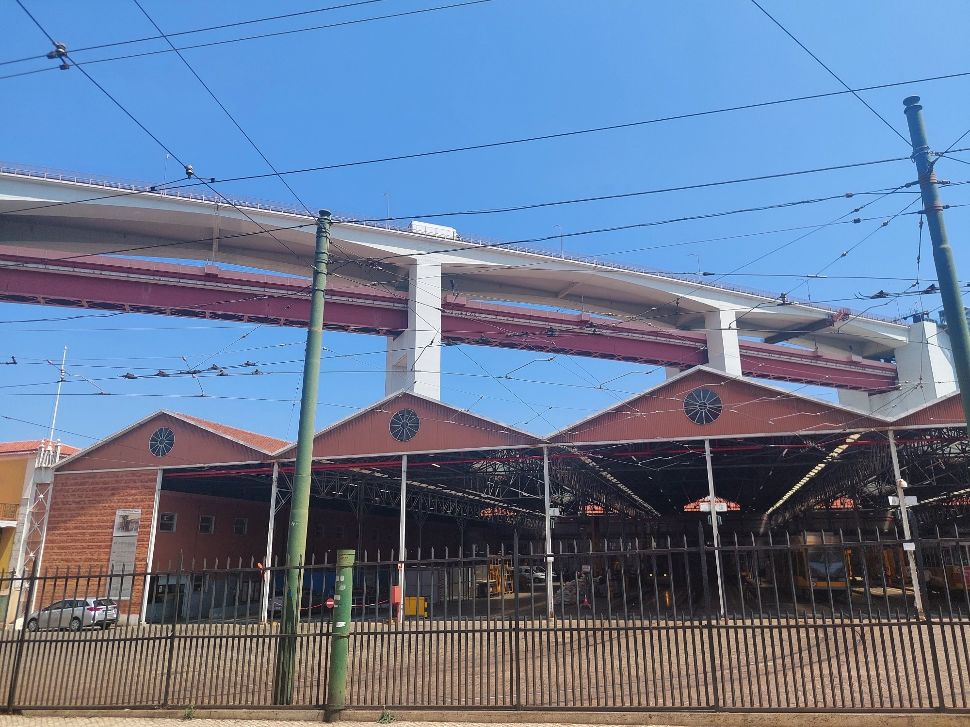 Zug/ Autobahnbrücke, darunter befinden sich offene Gebäudehallen. Vor dem Gebäude ist ein Zaun und es gibt zahlreiche Stromkabel.