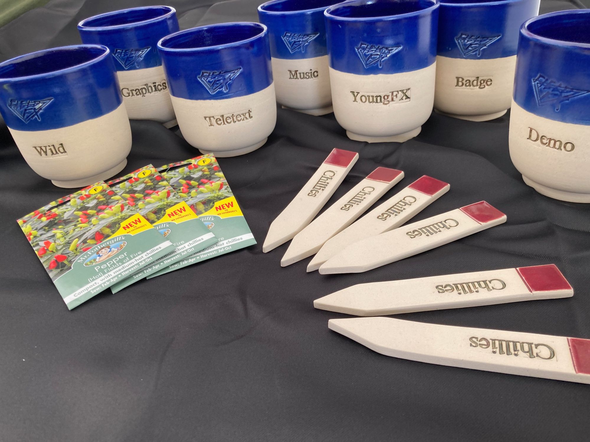 Seven pots, with blue tops and white bases, with Field-FX logos embossed and the words "wild" "graphics" "teletext" "music" "YoungFX" "Badge" "Demo" on them. In the foreground there are chilli seeds and matching "Chillies" plant labels made in the same pottery as the pots, with red tops.