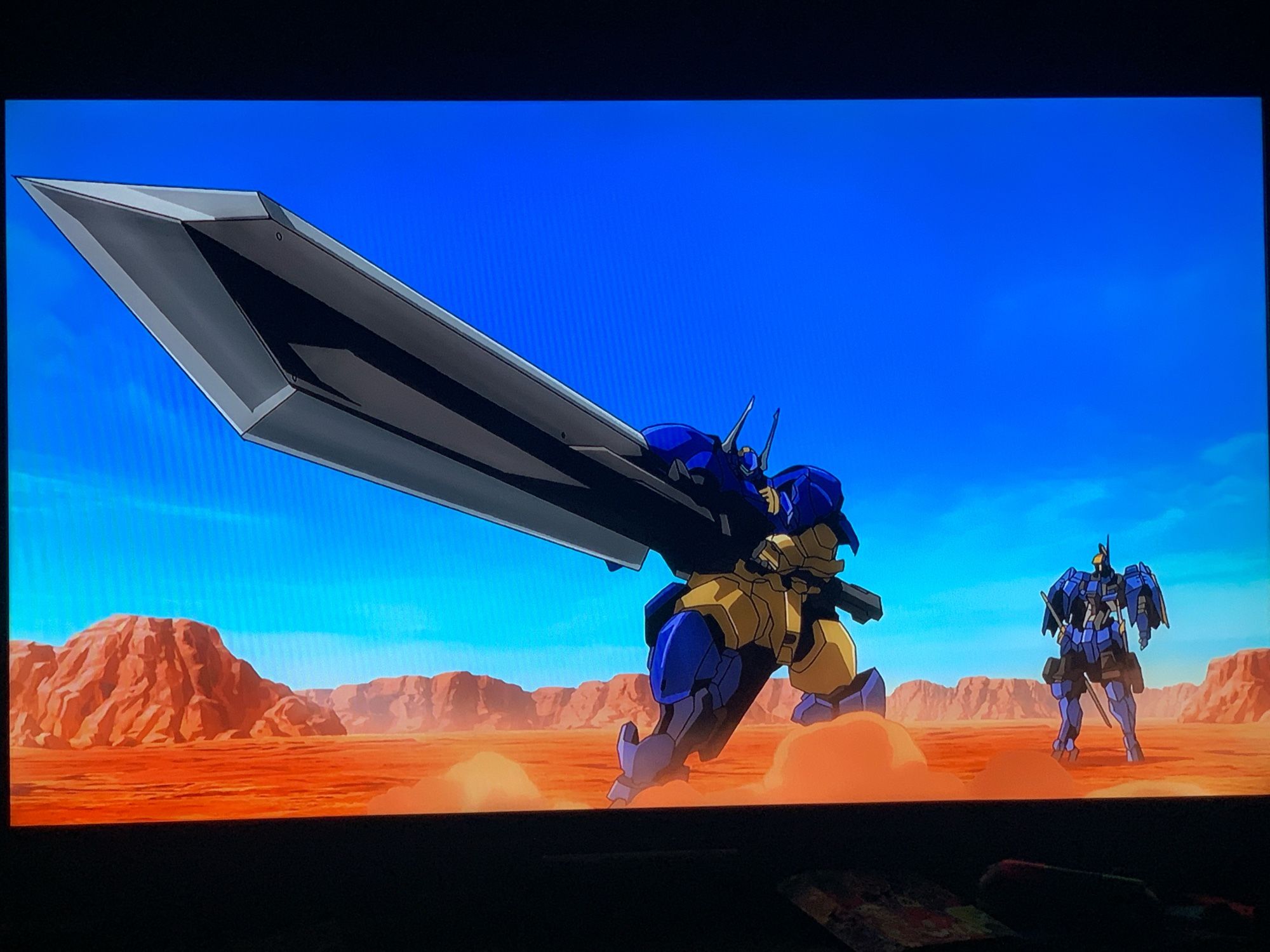 Shot from Mobile Suit Gundam: Iron-Blooded Orphans of two mechs on a desert surfacr, one's a thick boy holding a very long sword.