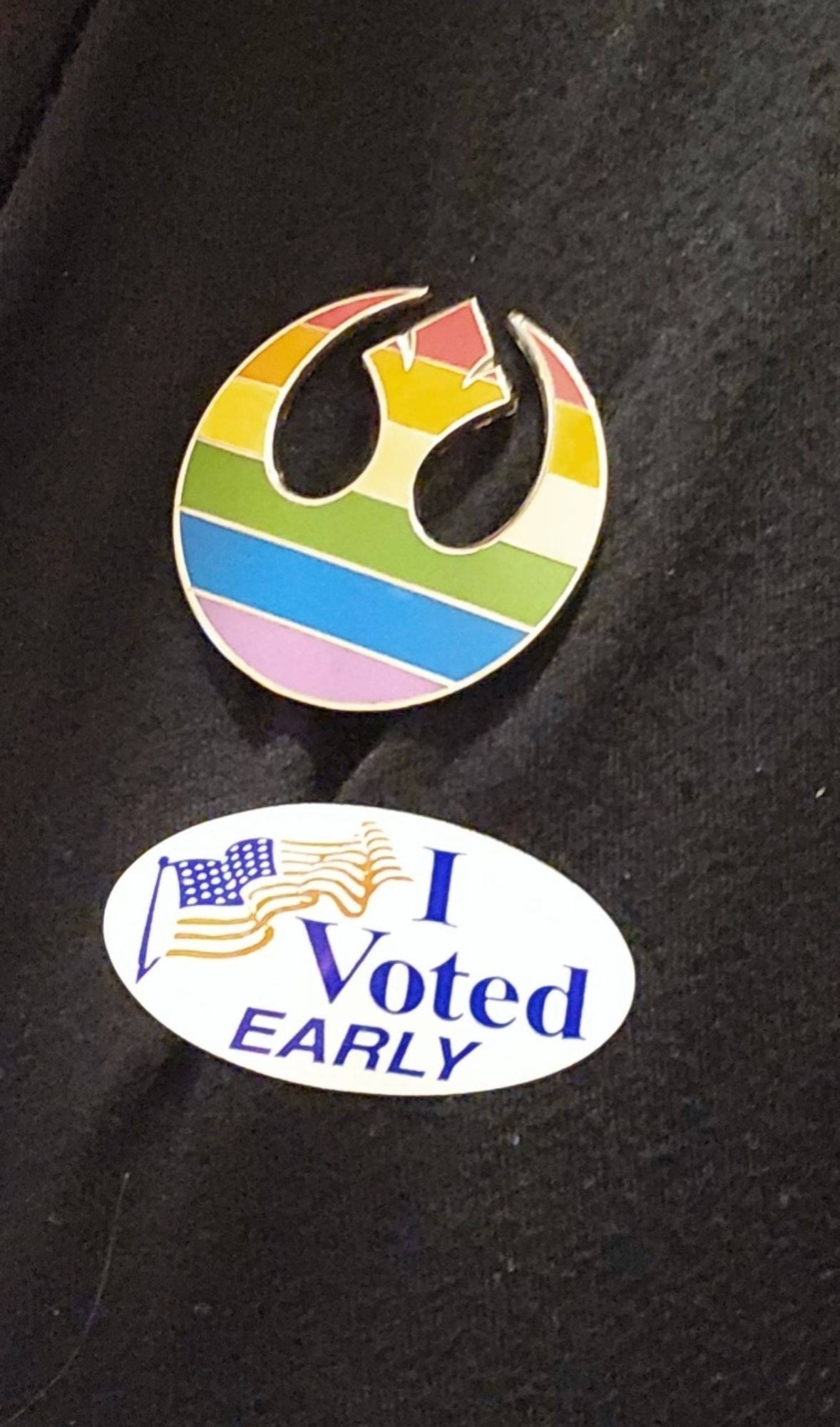 A picture of a white oval shaped sticker with an American flag illustration on it and the words I voted early in blue text. Above it is a e pin in the shape of the Rebel Alliance Firebird from Star wars. It is striped in a rainbow