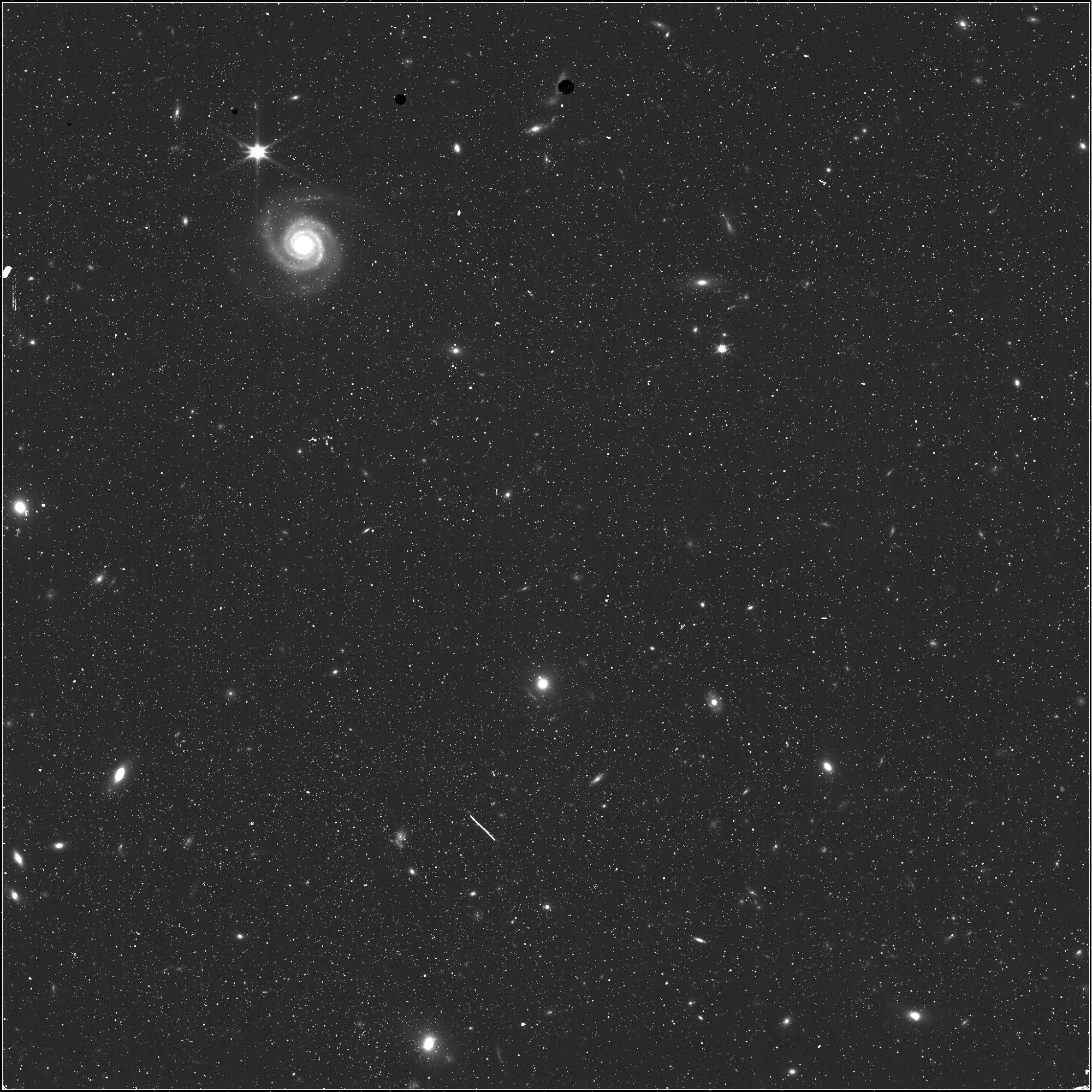 Black and white near-infrared image. The largest object is a galaxy on the top left. Smaller background galaxies also appear in the image, with a few stars in the Milky Way.