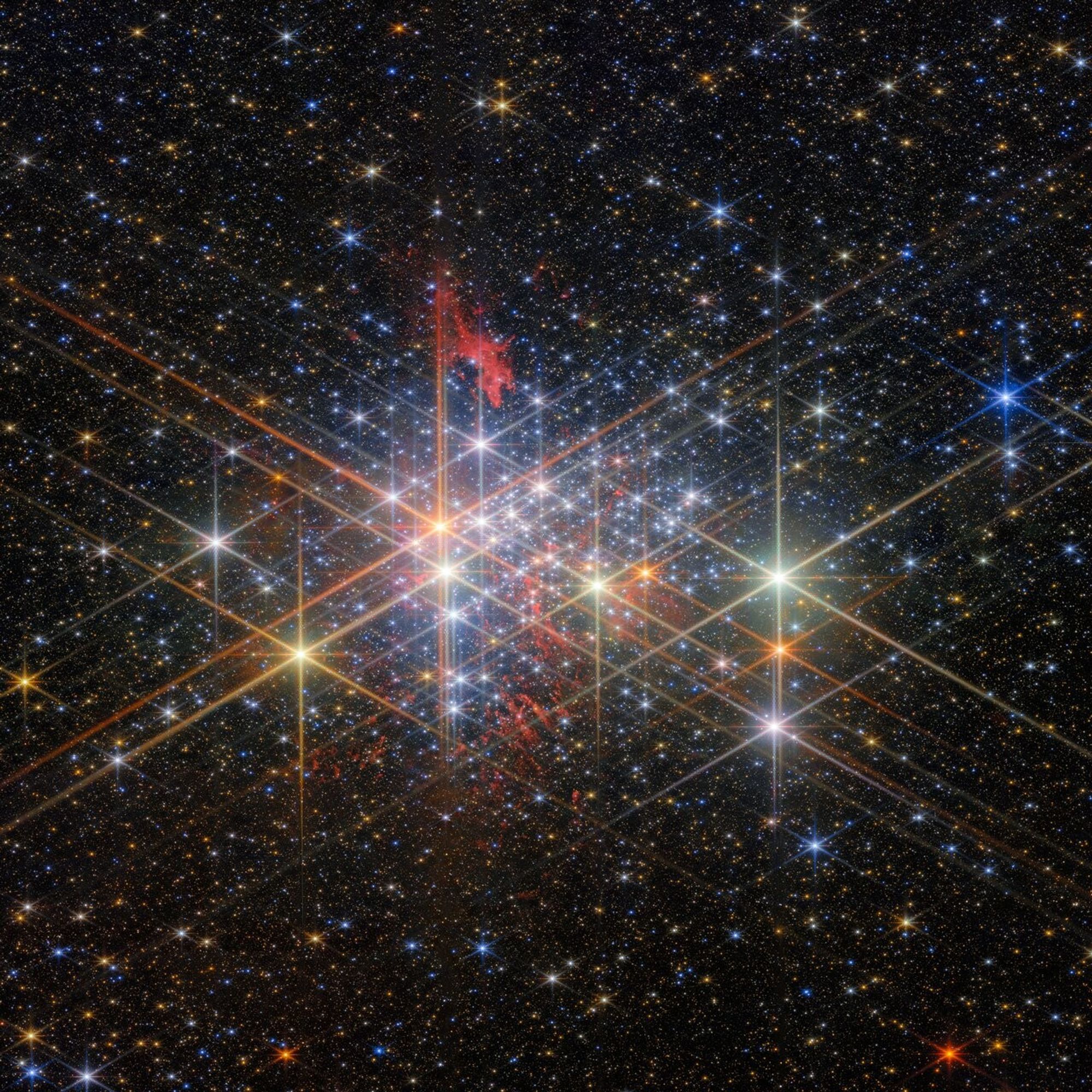 A dense cluster of bright stars, each with six large and two small diffraction spikes, due to the telescope’s optics. They have a variety of sizes depending on their brightness and distance from us in the cluster, and different colours reflecting different types of star. Patches of billowing red gas can be seen in and around the cluster, lit up by the stars. Small stars in the cluster blend into a background of distant stars and galaxies on black.