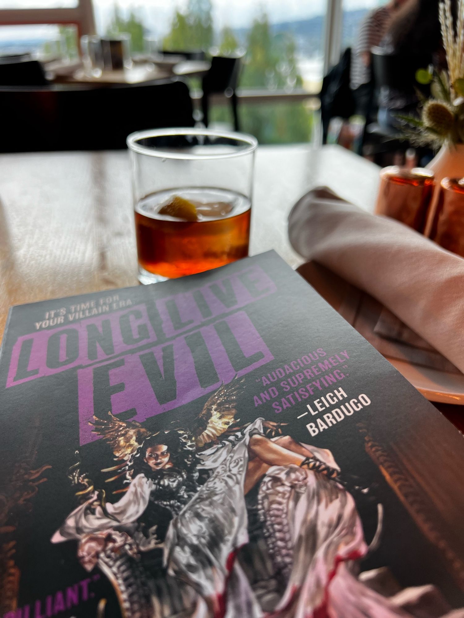 A copy of Long Live Evil on a table with an Old Fashioned in a rooftop restaurant.