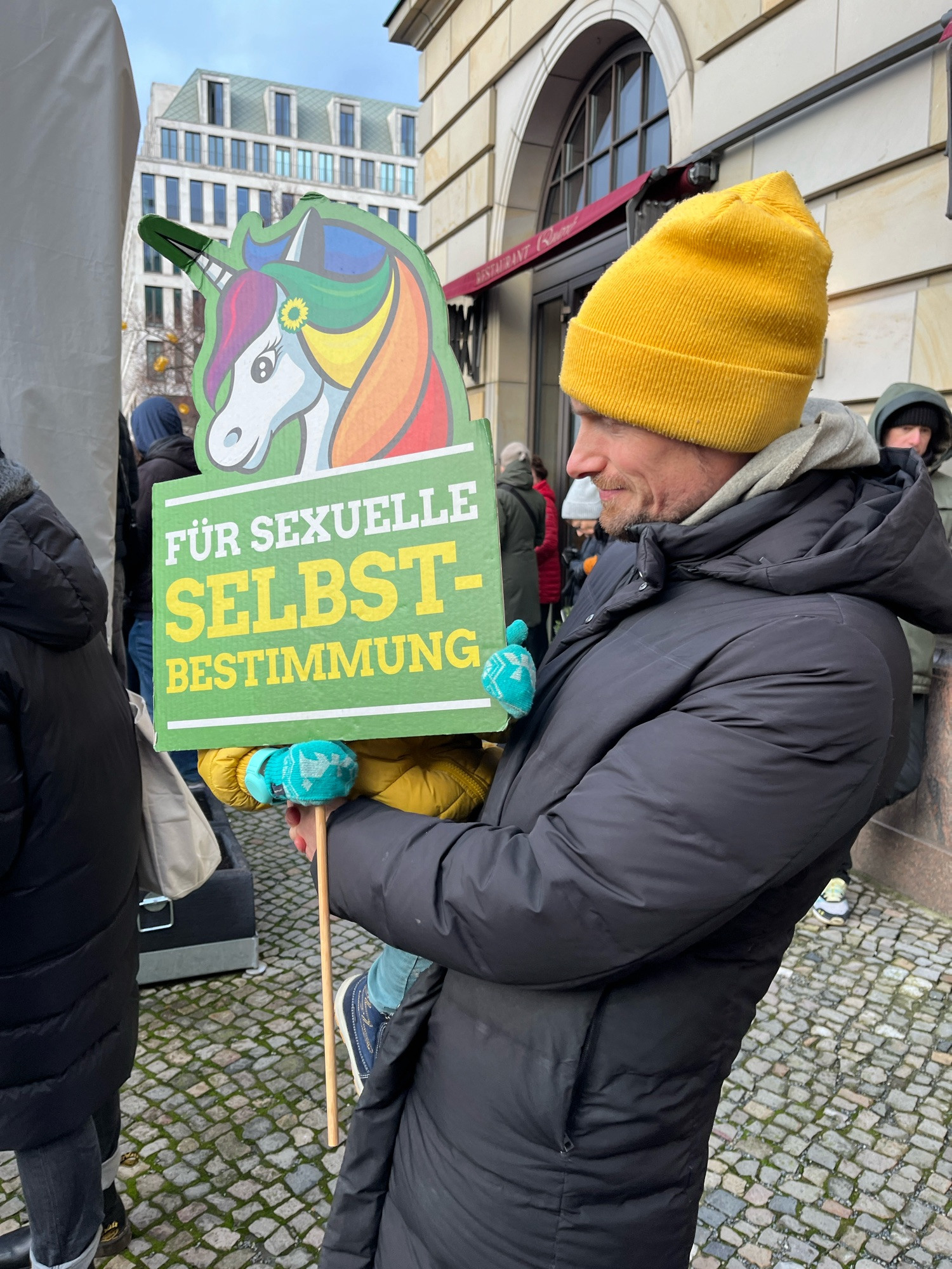 Ein Mensch mit gelber Mütze trägt sein Kind auf dem Arm, das wiederum ein Schild mit einem bunten Einhorn und der Aufschrift „für sexuelle Selbstbestimmung“ vor sich hält.