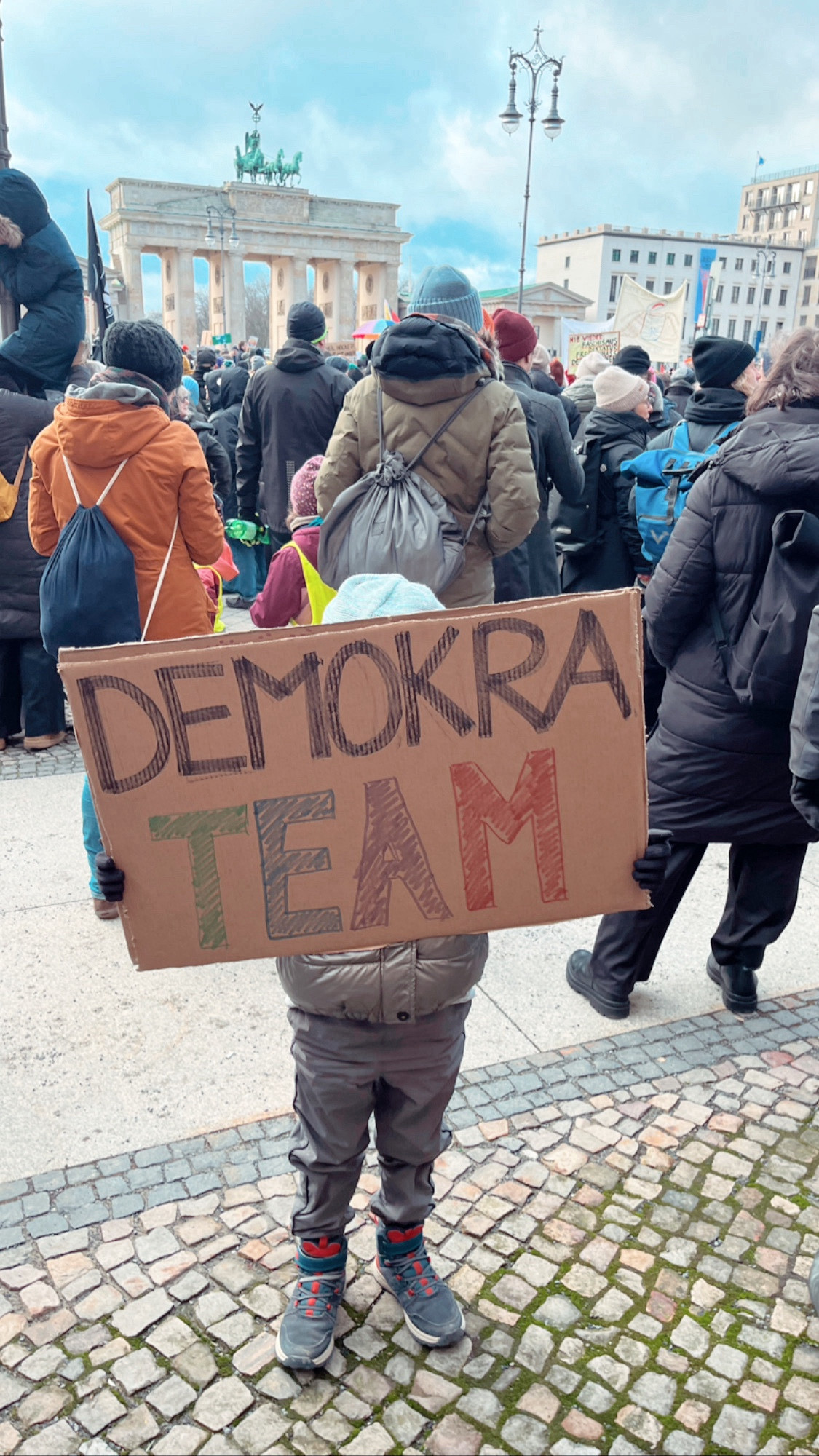 Kind hält ein Schild mit der Aufschrift Demokrateam, im Hintergrund viele Menschen und das Brandenburger Tor.