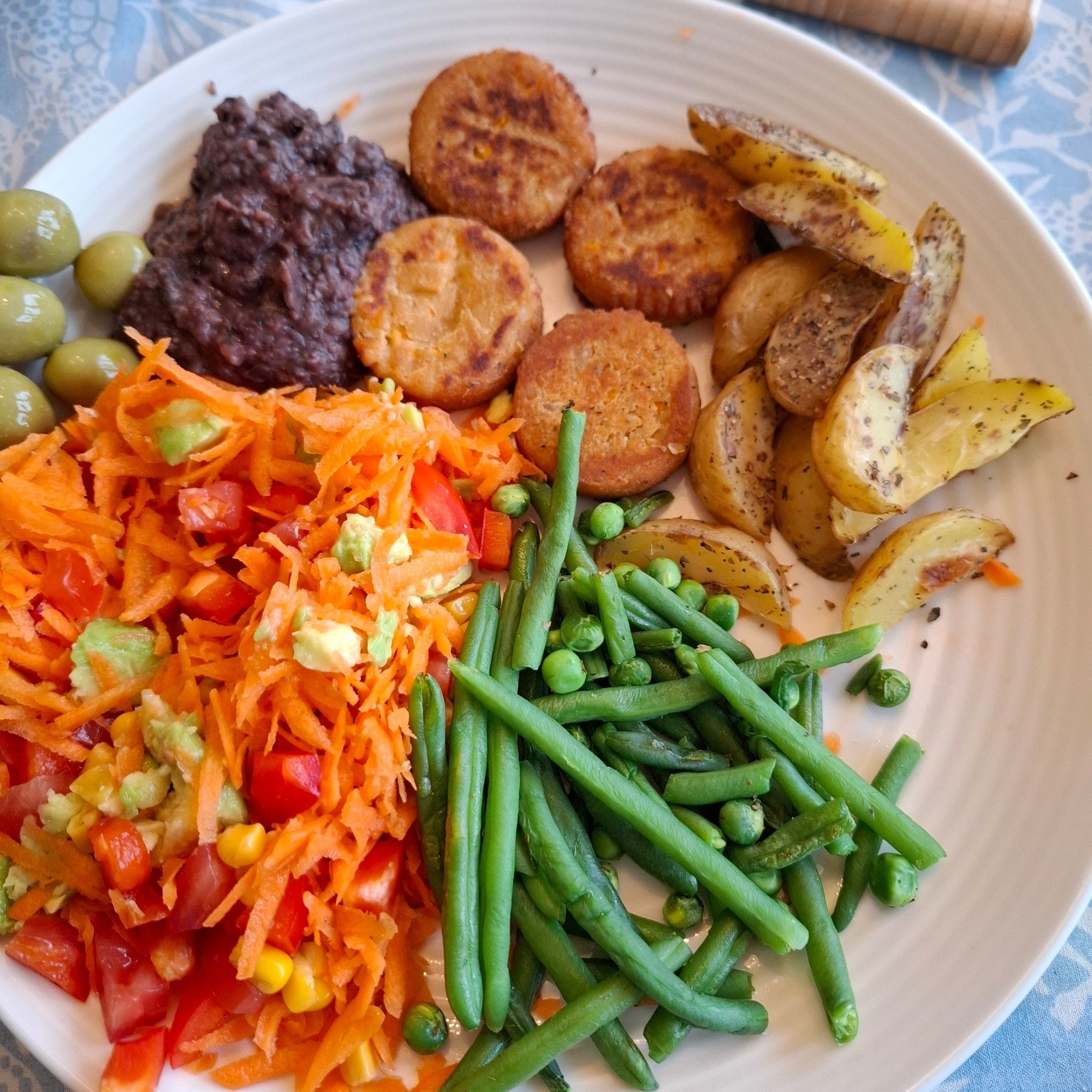 Oliven, bønnepuré, falafler, stekte potetbåter, erter, aspargesbønner, erter og råkost med gulrot, litt avocado, mais, tomat og paprika på en hvit tallerken