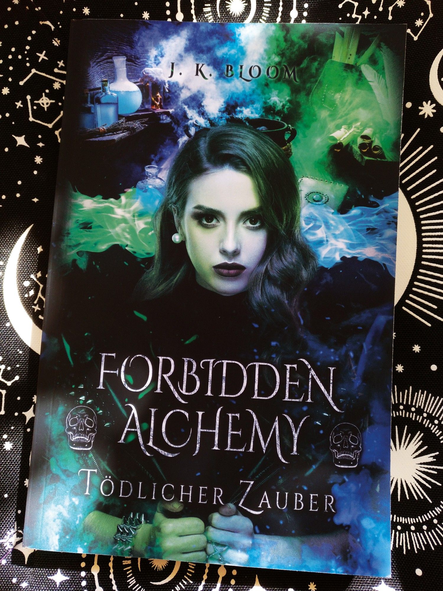 Das Cover des Buchs "Forbidden Alchemy - Tödlicher Zauber" der Autorin J. K. Bloom. Darauf ist die Protagonistin Cara Hartley zu sehn und im Hintergrund Zaubertränke, Kessel, etc.