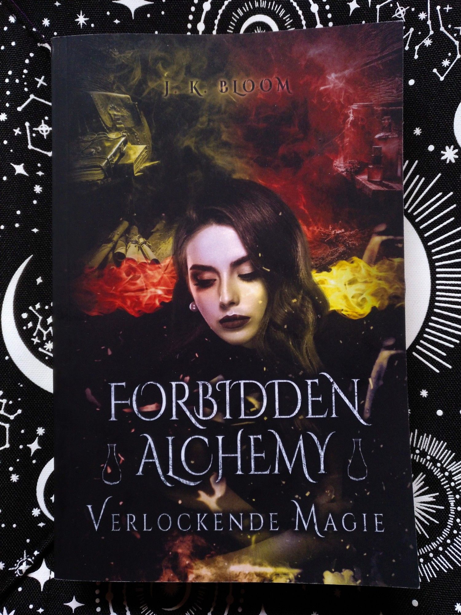 Das Cover des Buchs "Forbidden Alchemy - Verlockende Magie" der Autorin J. K. Bloom. Darauf ist die Protagonistin Cara Hartley zu sehen und im Hintergrund verschiedene Bücher, Schriftrollen, Tränke, etc.