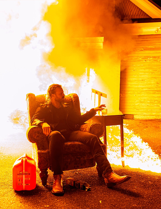 Hangman Page sitting on a chair while a house burns behind him