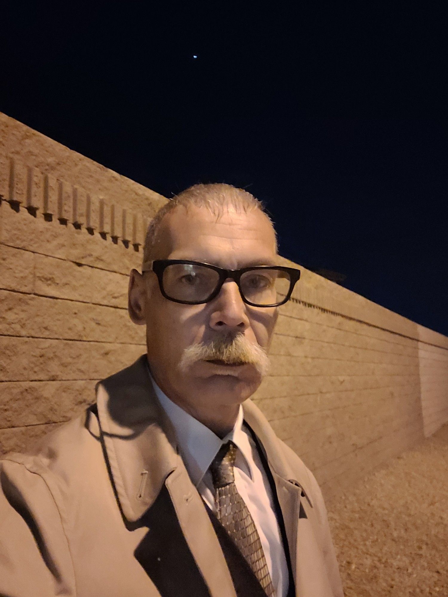 A devilish handsome older man with glasses, a 70s pornstash and a caesar haircut wearing a shirt and tie with an overcoat.