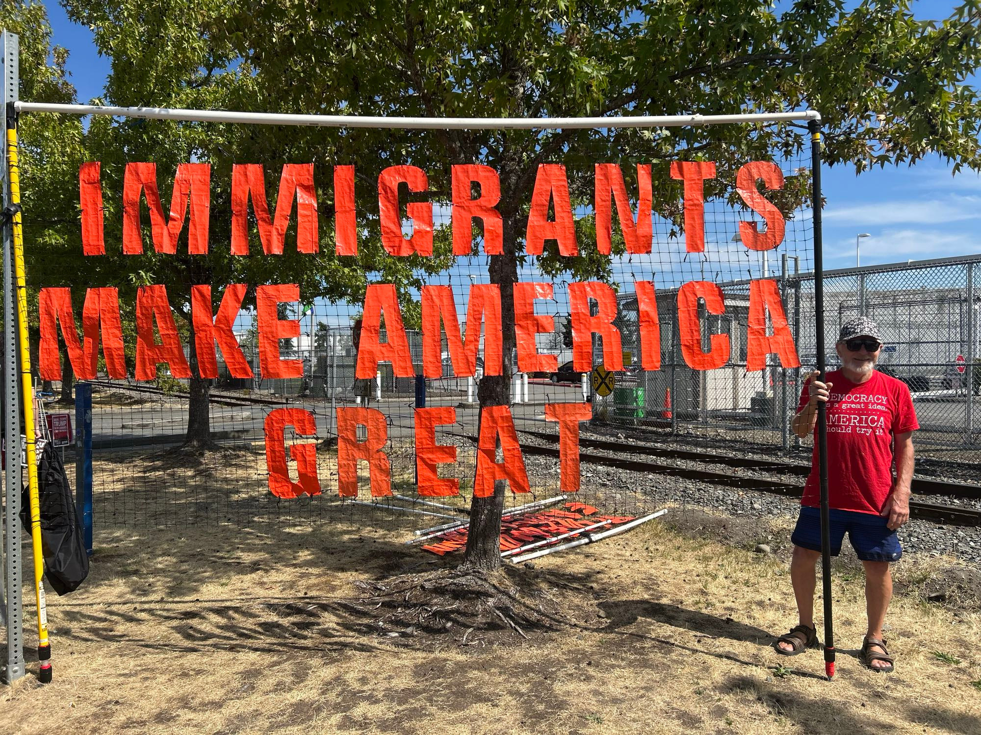 A man in a red shirt holds a banner reading, “Immigrants make America great.”
