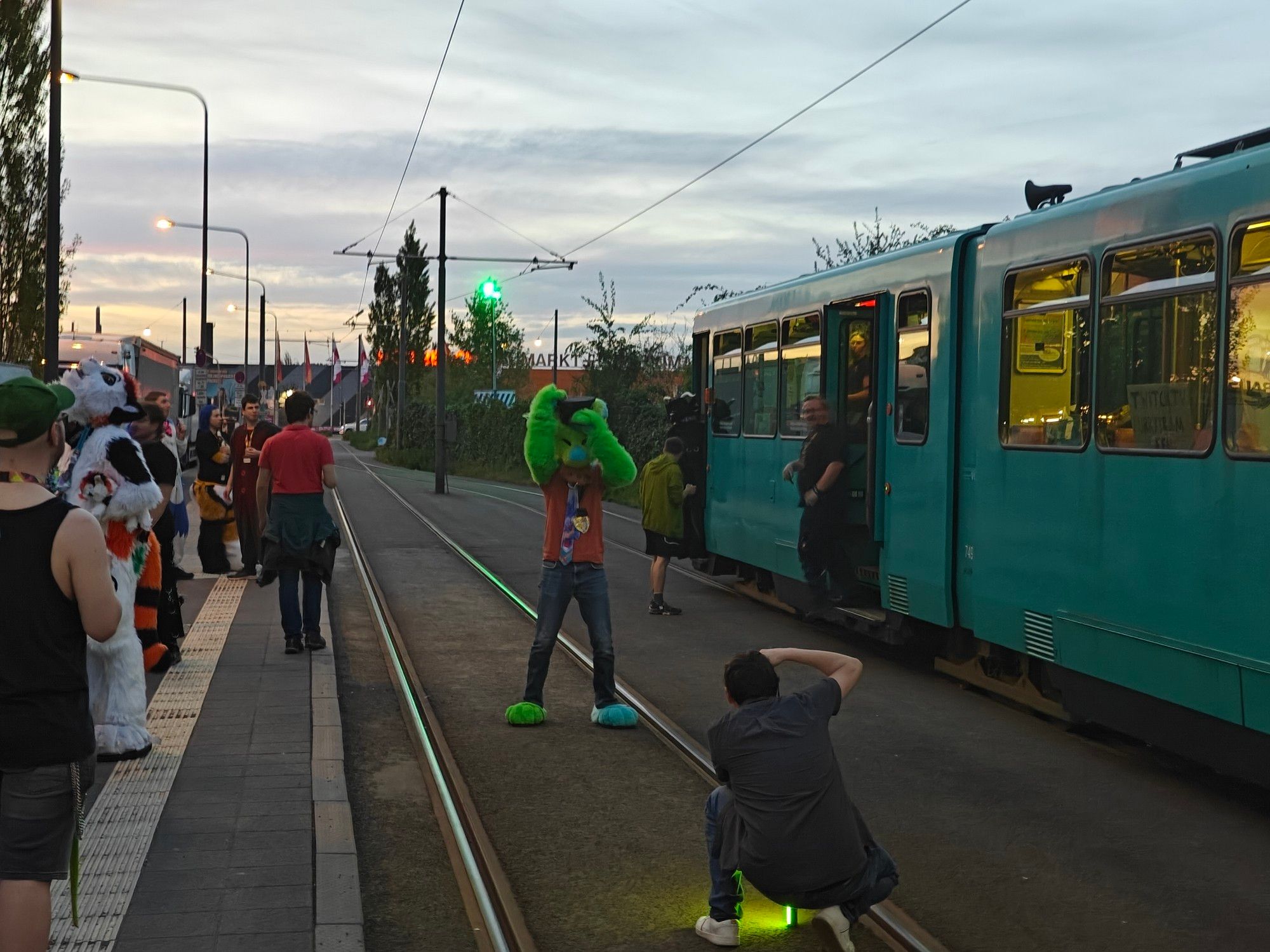 A picture of the tram and the attendees of the Frankfurt Furry Tram enjoying a break at a tram stop. It's evening, the sun has already set and we see an orange hue at the horizon.
Several Suiters have disembarked the tram and enjoy the evening cool.
One is having a picture taken of them in the middle of the tram tracks.