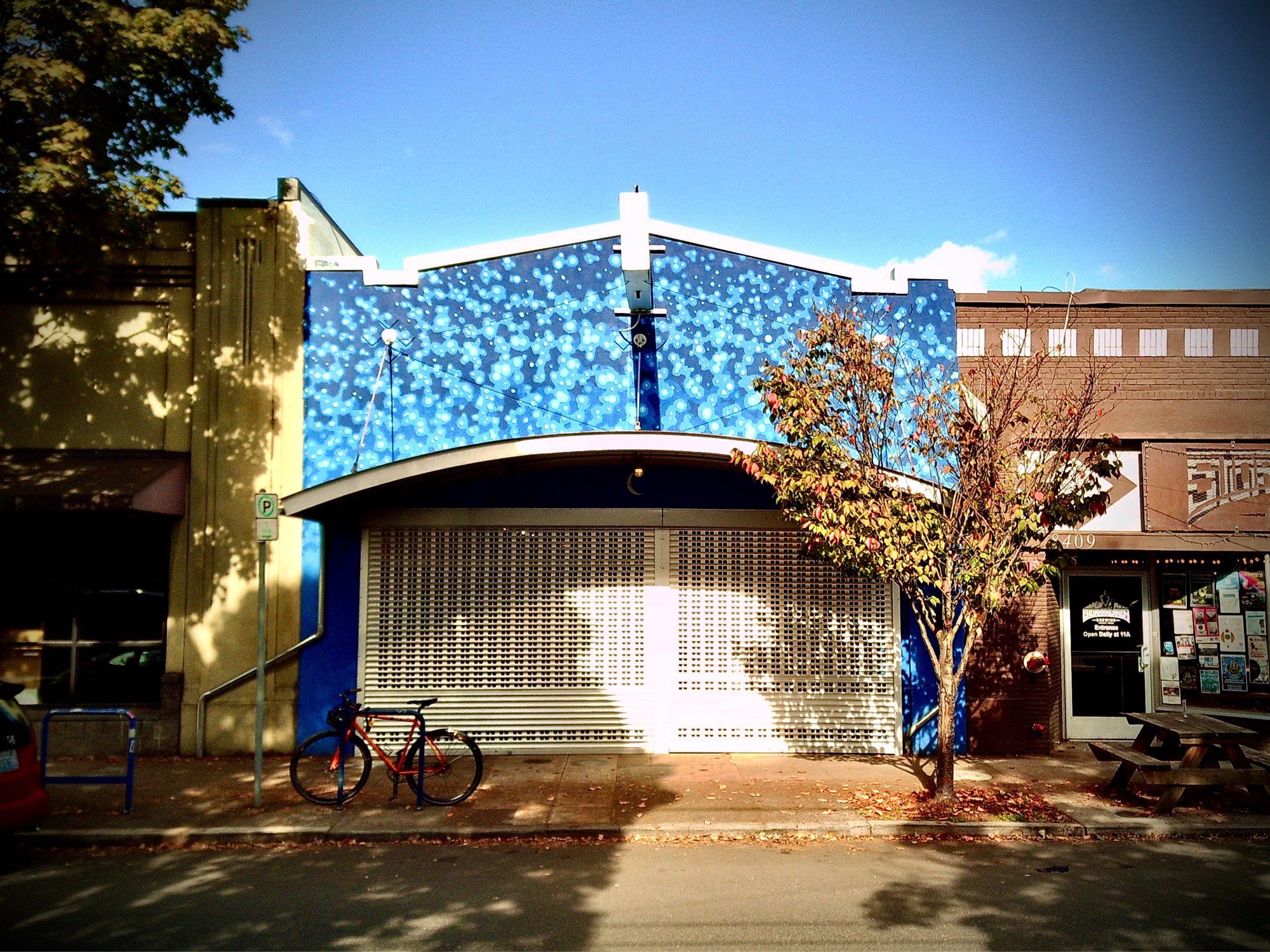 Looking across a street at a blue-painted storefront, the upper half dappled in different shades, its broad, white-painted gate shuttered.