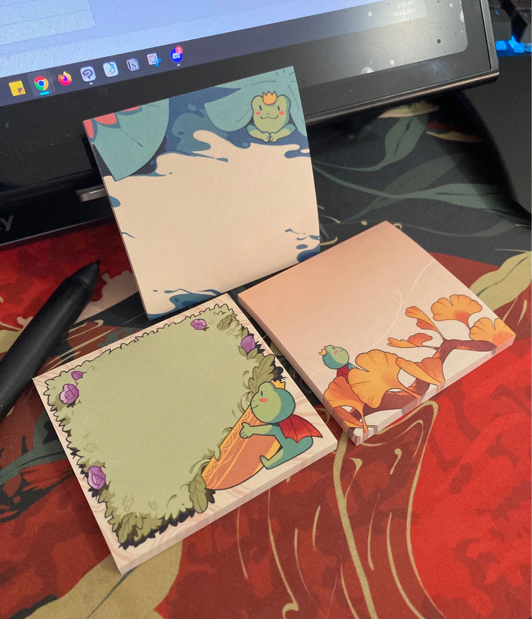 A photo of three sticky note pads with drawings of a frog in various poses. One under a lily pad, holding a flower pot, and one sitting on a ginkgo plant.