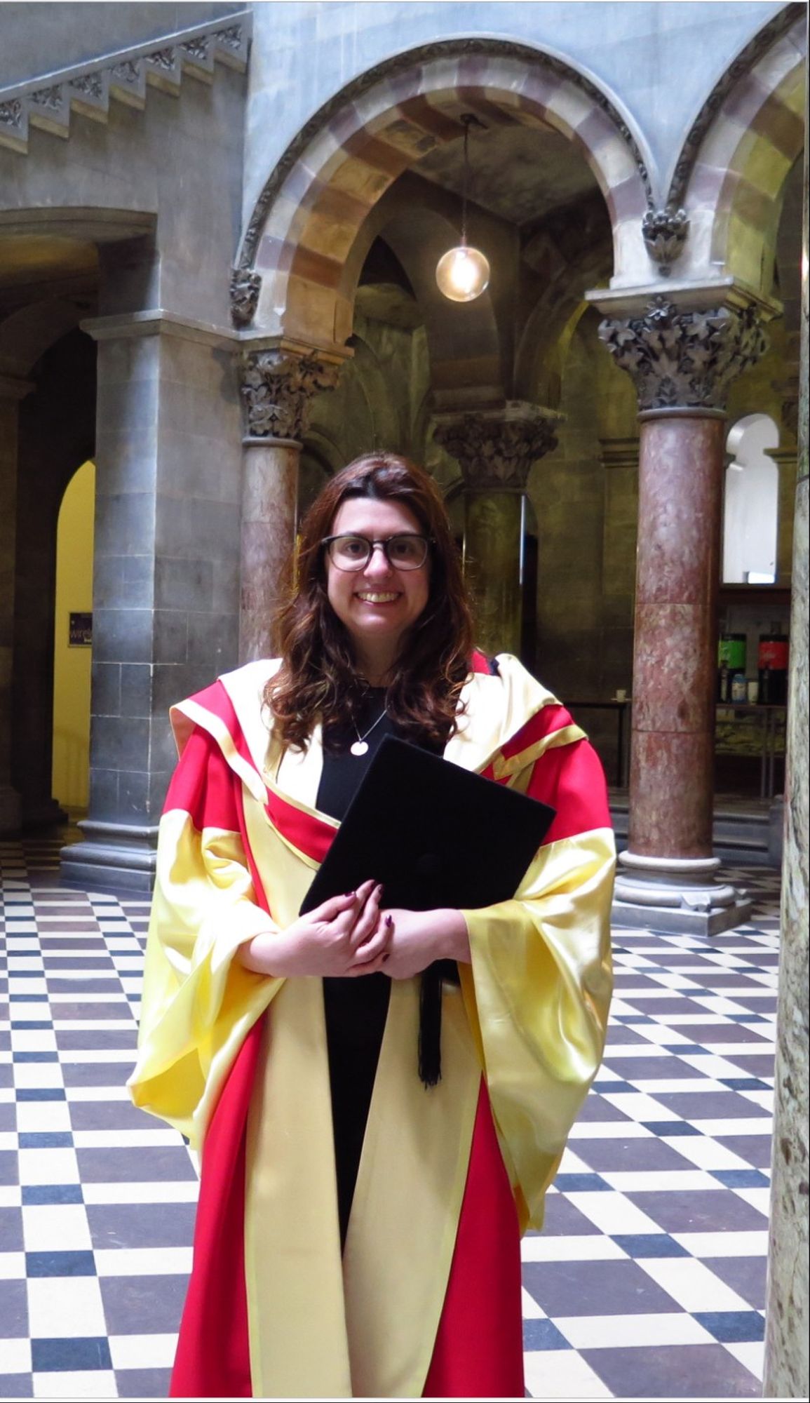 A young white woman with long dark hair and glasses, wearing a yellow and red robe and holding a mortarboard, and grinning.