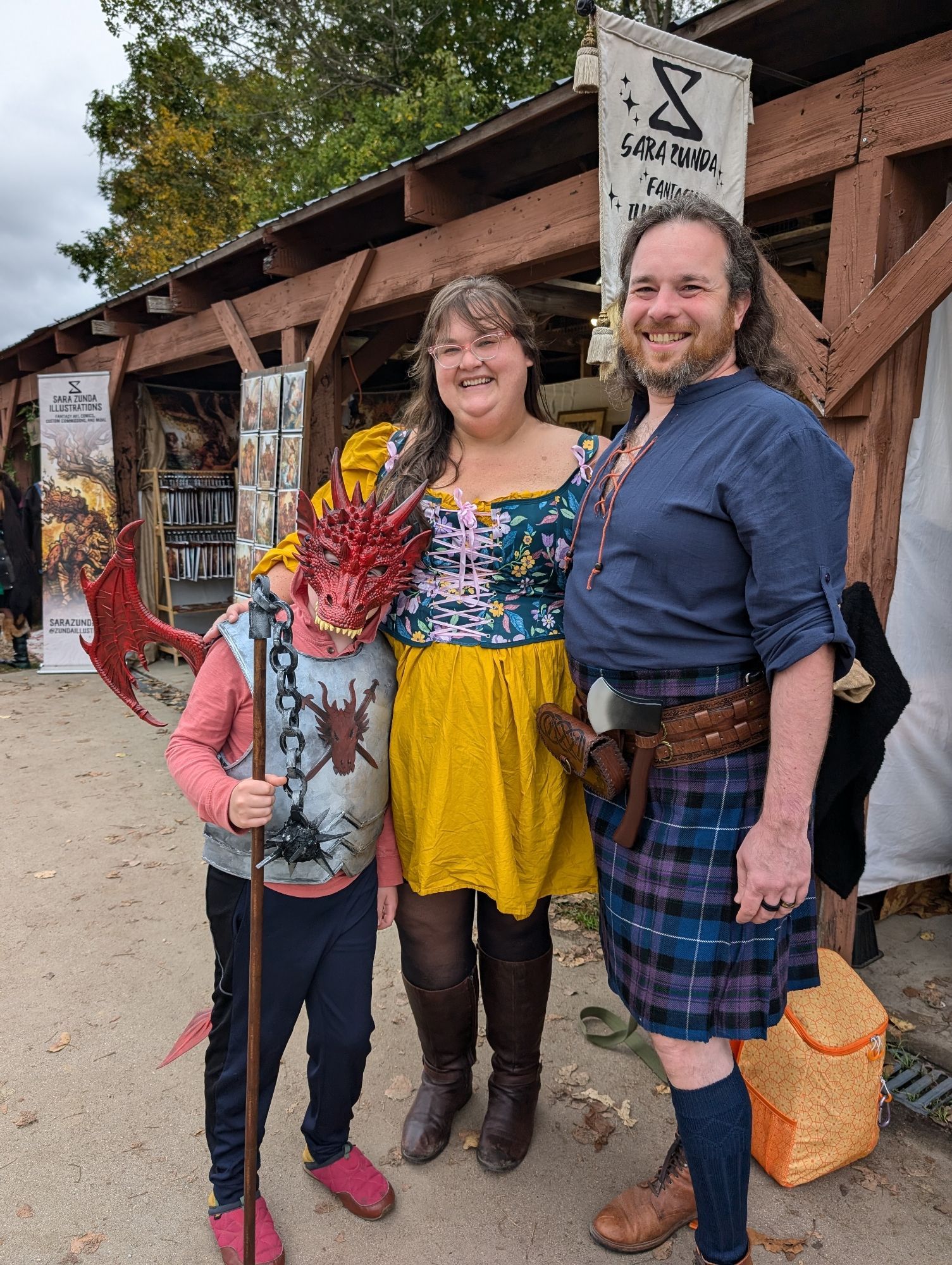 An almost 10 year old child at a Renaissance Faire wears metallic-looking armor crafted from foam, with added dragon wings and mask, and holds a flail also made from painted foam. Their parents, a white woman in a corsette and a white man in a kilt, pose alongside.