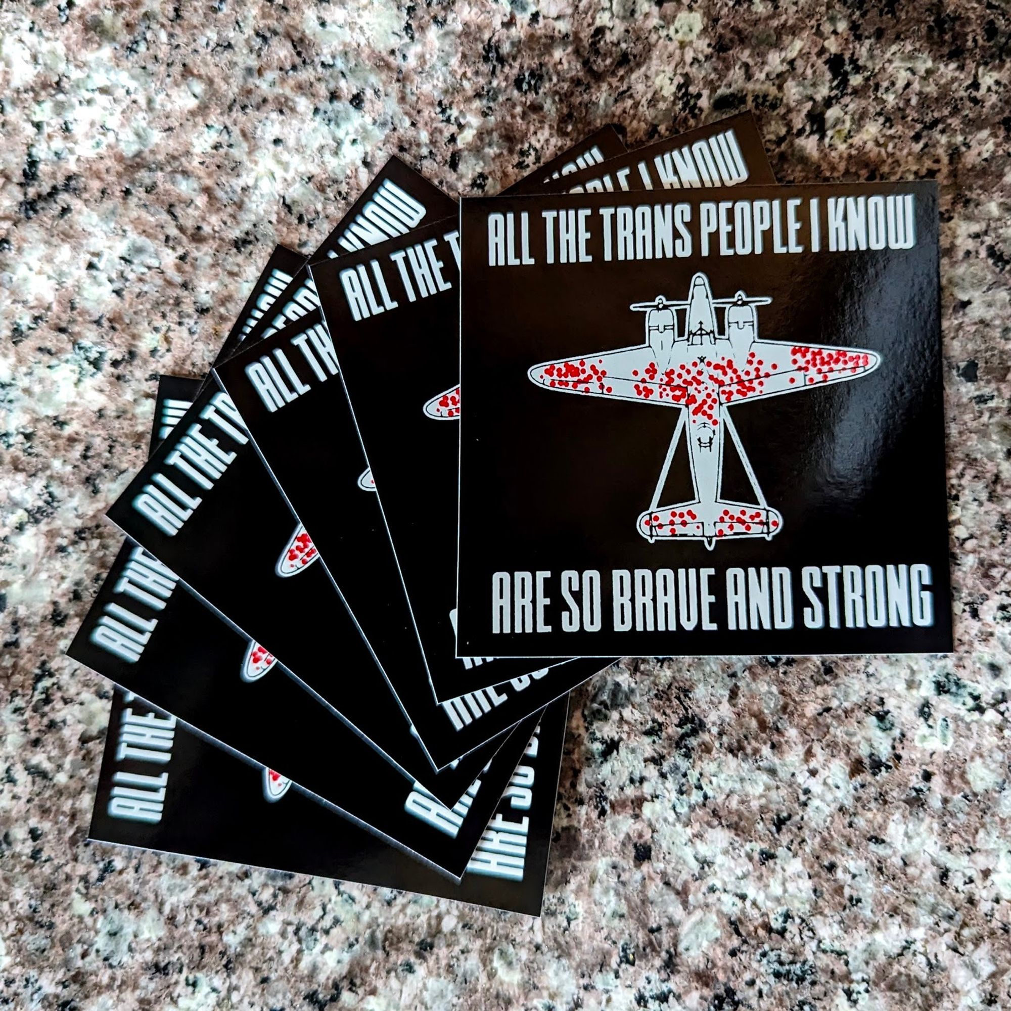 A fan of stickers, each is square with "All the trans people I know are so brave and strong" split between top and bottom around a diagram of a WW2 plane with damage markers for planes that made it back home