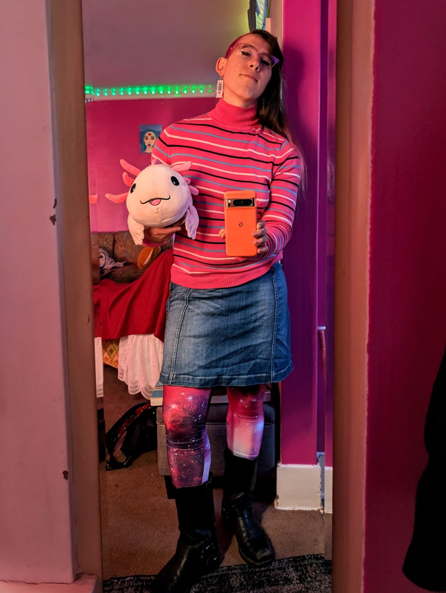 Me in the mirror holding a big stuffed pink axolotl. I'm wearing a stripey pink sweater. jean skirt, and starry pink tights with tall black boots. The walls behind me are pink af