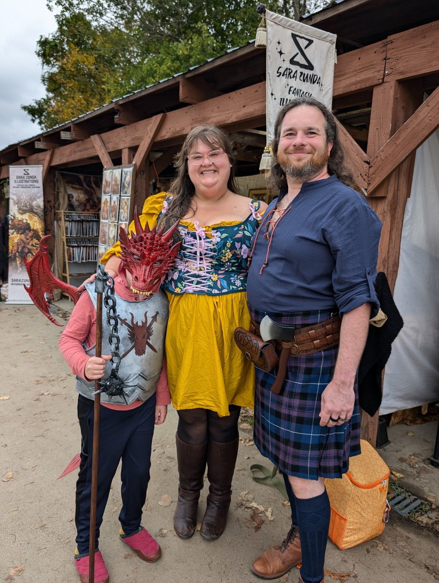 Long-haired bearded man in a kilt, long-haired woman with glasses in a yellow dress and floral bodice and boots, and a child in a dragon mask and armor with wings holding a staff with a morningstar on the end