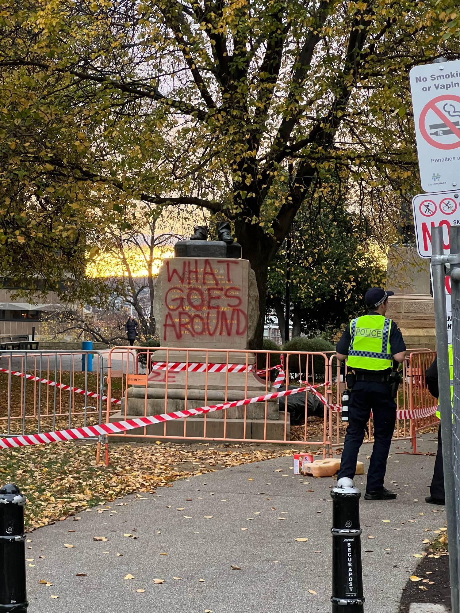 A stone plinth in a city park with the feet of a statue on top. The statue has been cut off and lies on the ground behind the plinth. A police officer is in the middle ground.