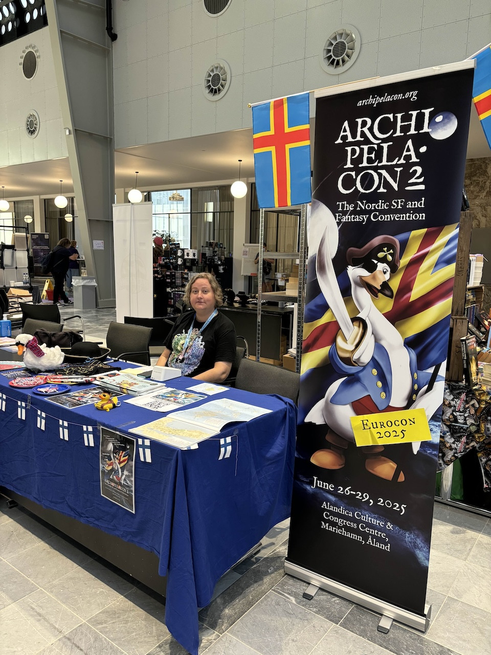 A woman sits at a convention table decorated with a blue tablecloth featuring Finnish flags. The table displays various promotional materials and merchandise related to Archipelacon 2, a Nordic science fiction and fantasy convention. A large banner beside her features pirate swan-themed artwork and the Åland flag.