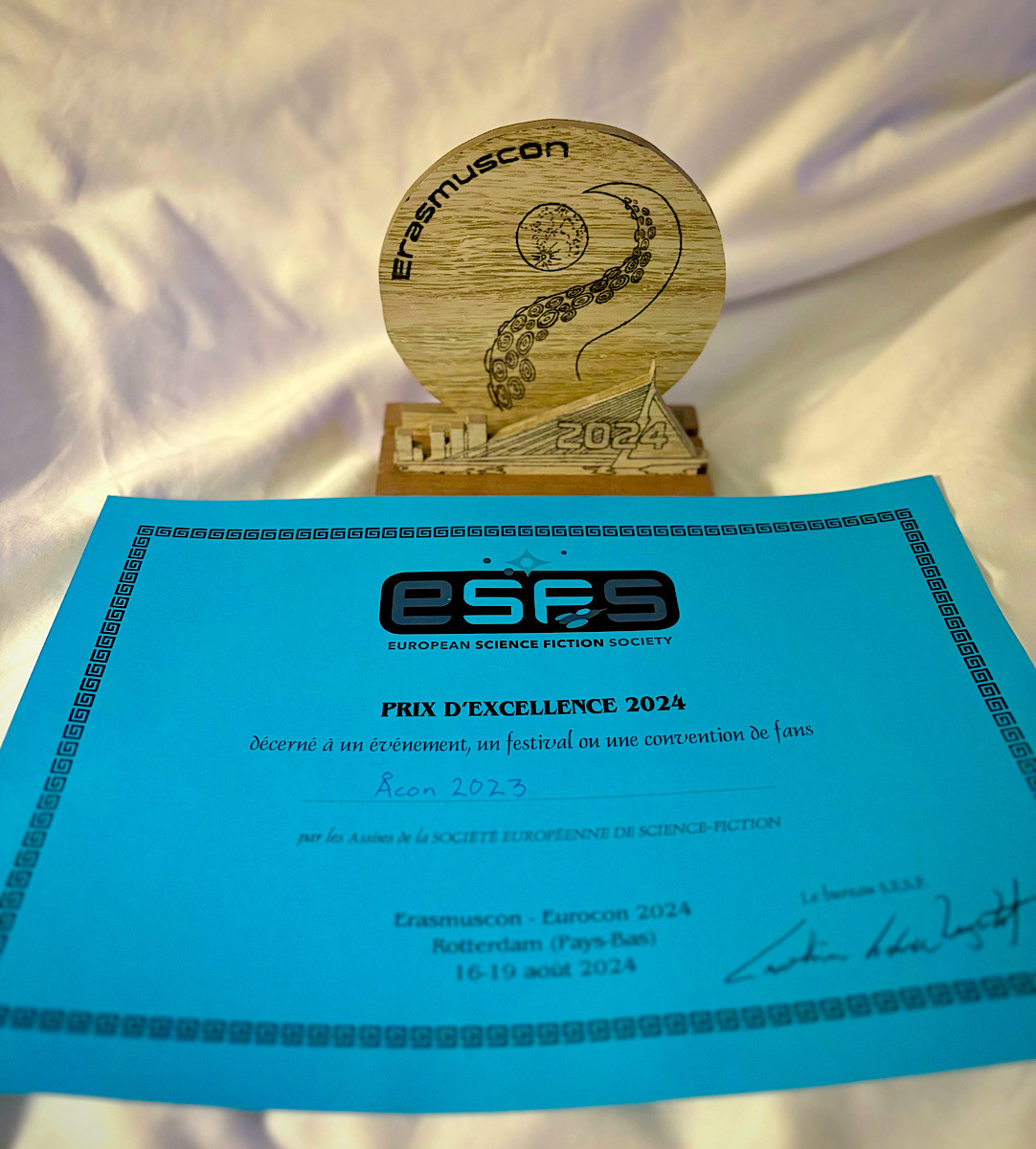 A wooden award trophy and a certificate. The trophy is engraved with “Erasmuscon”, “2024”, and images of a tentacle and a planet. The blue certificate, issued by the European Science Fiction Society is the prix d’excellence 2024 for Åcon 2023, for best science fiction event of the year.