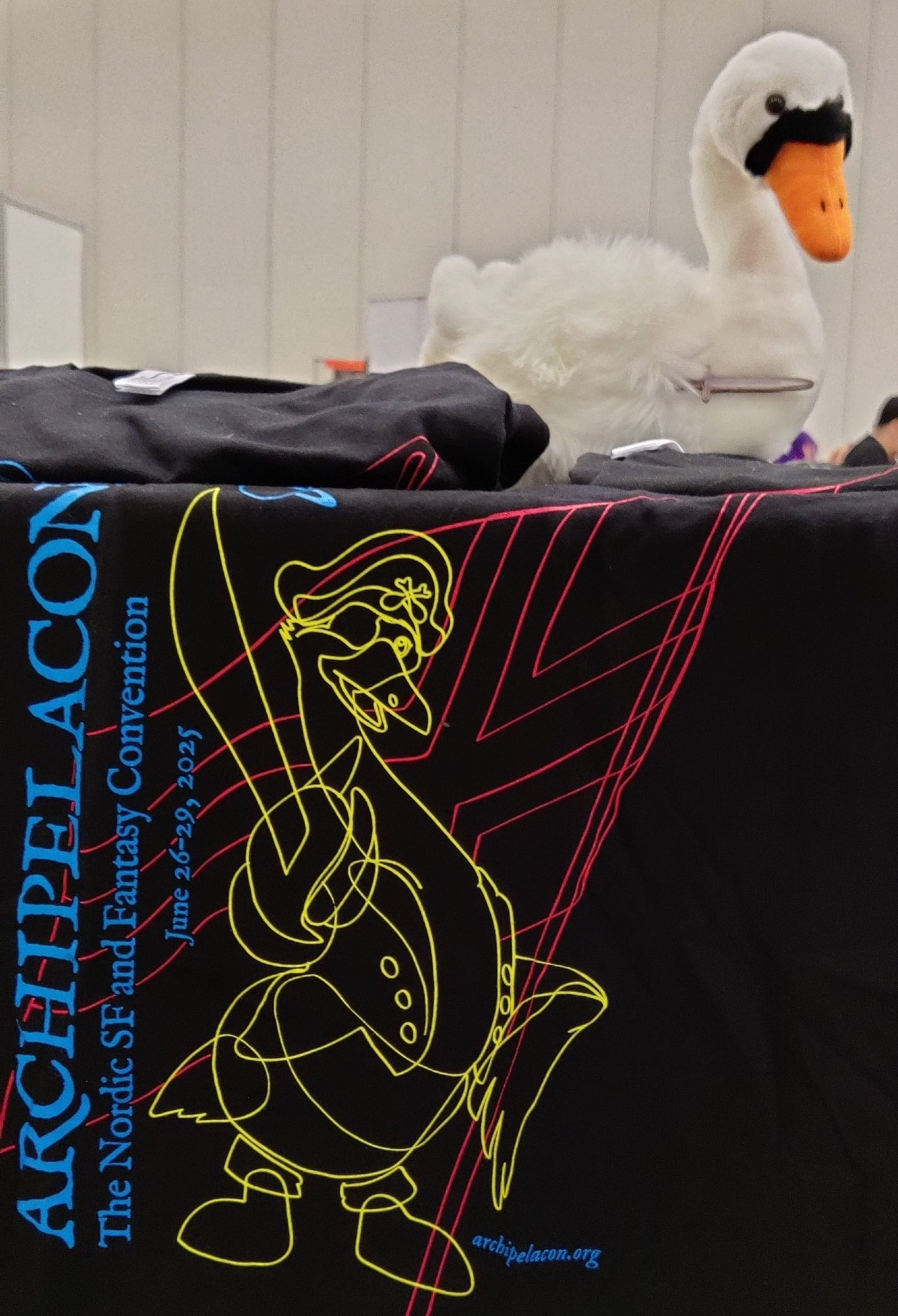 A T-shirt hanging from a table, with the text “ARCHIPELACON – The Nordic SF and Fantasy Convention – June 26–29, 2025” and an image of a cartoon duck wearing a pirate hat; the design is neon colors on a black shirt. A white swan plushie sits on the table.