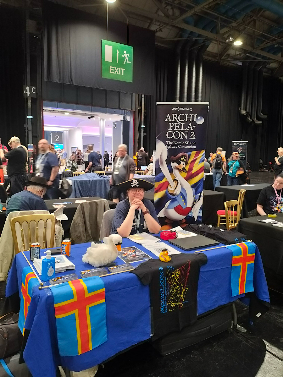 A person sitting at a table draped with colorful flags and promotional materials for Archipelacon 2, a Nordic science fiction and fantasy convention. The table also has a plush seagull and beverage cans. People are milling about in the background.