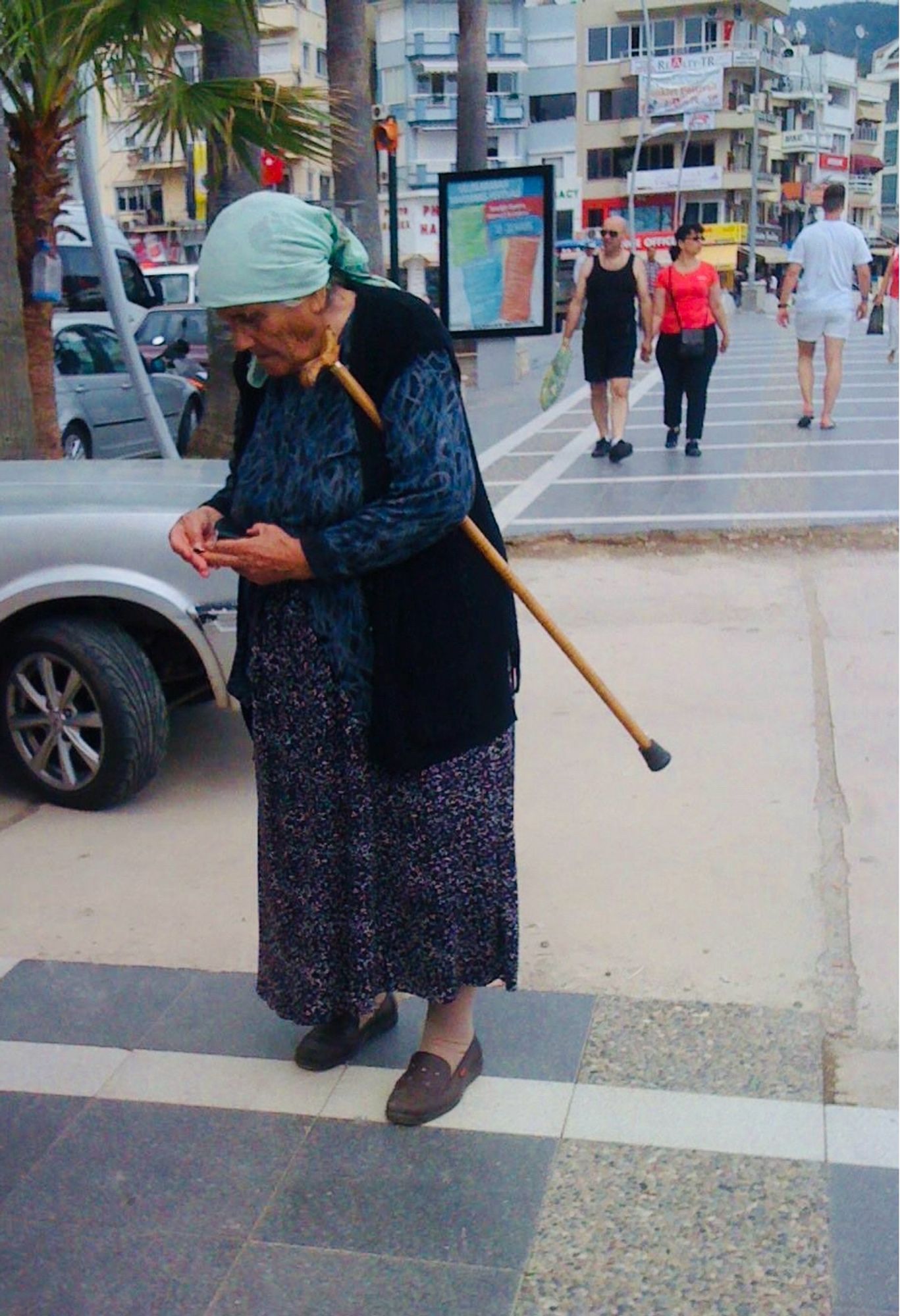 An old Turkish woman pauses on the promenade with her walking stick tucked up under her arm. She is dressed in a shapeless dark-blue ankle-length cotton dress, a long black unbuttoned sweater vest, and a light-blue headscarf over her hair. In the background, young people stroll in tank tops and shorts on the paved along the waterfront, before the summer tourists arrive in the resort. Marmaris, Türkiye 🇹🇷 🌴(May 2012)