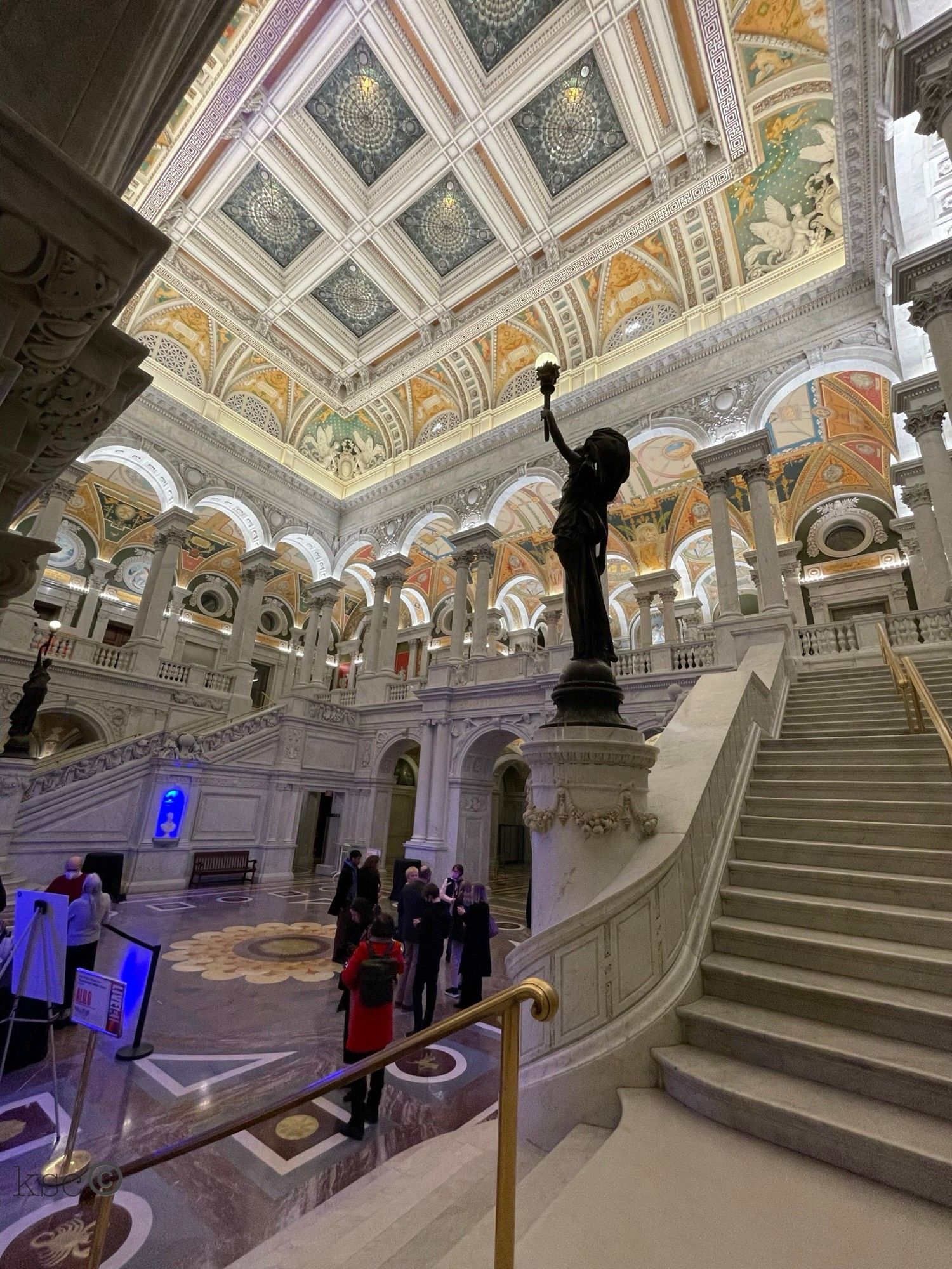 Photo of the Library of Congress interior shows a high vaulted ceiling decorated with paintings; dark sculpted light-bearers on plinths: and white-marble steps leading up to an arched colonnade that overlooks the large front-hall space. The Library is open to the public on Thursday evenings for musical performances and other events. Washington, DC (September 2022)