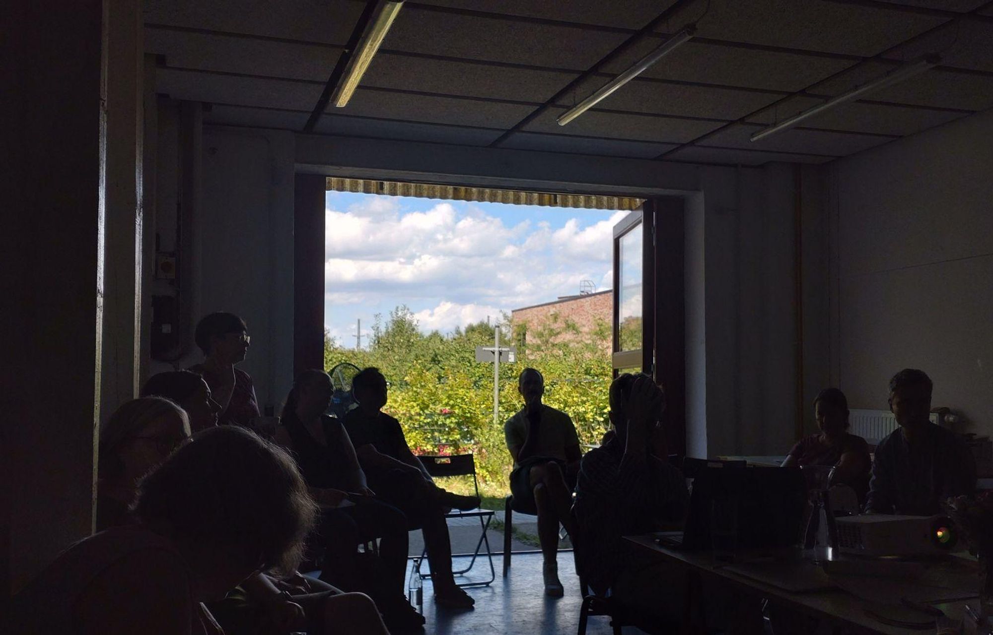 Großes Fenster mit Blick auf einen blau-bewölkten Himmel, viel Grün und eine Ecke Ziegelmauer des Gymnasiums Dresden Pieschen. Im Vordergrund Personen im Gespräch.