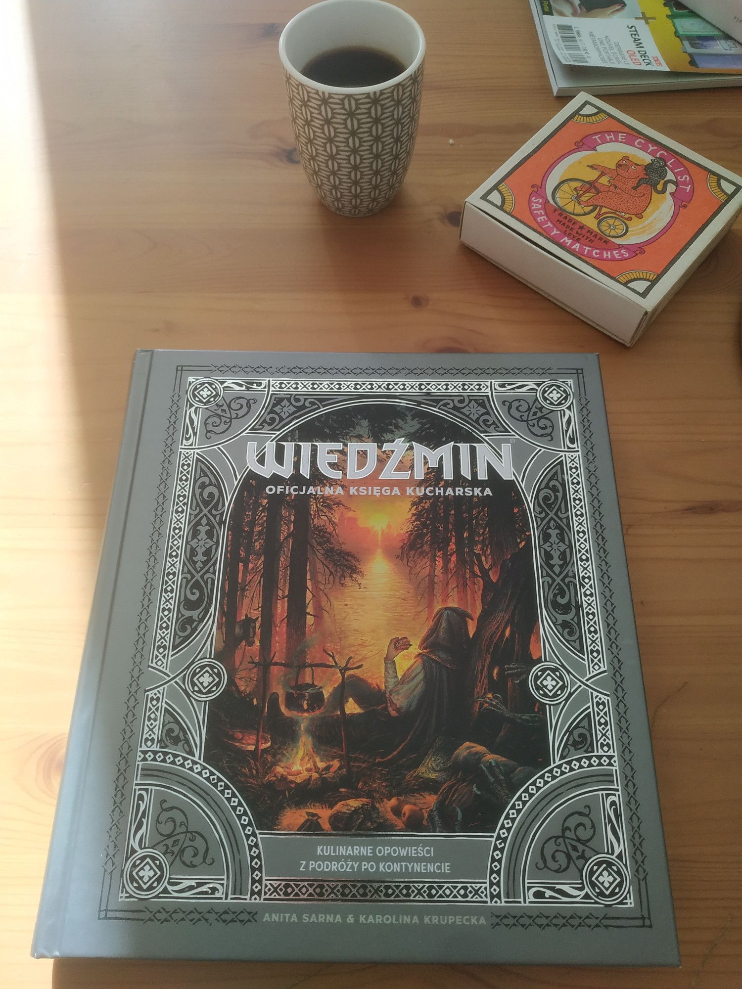 Livre de recette "Wiedzmin" (The Witcher en polonais), fermé, couverture visible. Couverture cadre argenté,  posé sur une table de cuisine en faux hêtre, à côté d'une tasse de café et d'une boîte d'allumettes orange avec un tigre sur bicyclette sur l'illustration. Un coin d'un magazine Canard PC Hardware en haut à droite. Un rayon de soleil sur la gauche de l'image