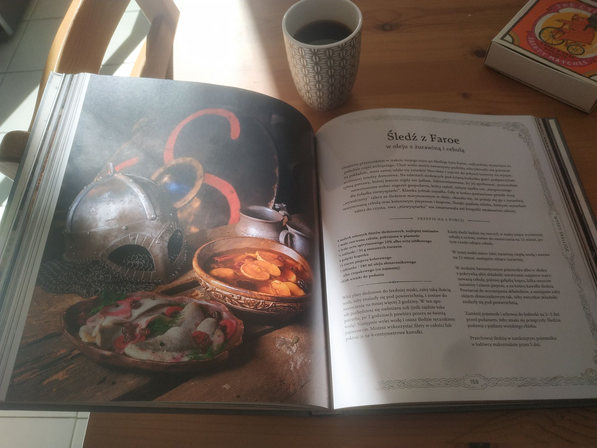 Livre de recette "Wiedzmin" (The Witcher, en polonais) ouvert sur la page d'une recette aux harengs, illustration page gauche du livre, recette à droite. La photo page gauche montre un bol de harengs, posé à côté d'un casque médieval et d'un bouclier en arrière-plan. Le livre de recettes est posé sur une table de cuisine en faux hêtre, à côté d'une tasse de café et d'une boîte d'allumettes orange avec un tigre sur bicyclette sur l'illustration. Un rayon de soleil touche la page gauche du livre.