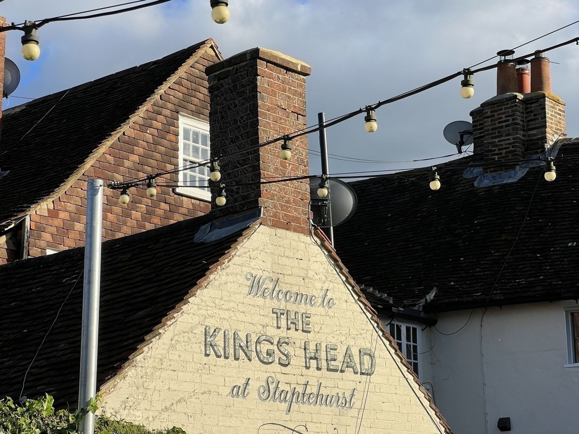 A pub with a sign that reads "Welcome to The Kings Head at Staplehurst" is beneath a string of bulb lights against a partly cloudy sky.