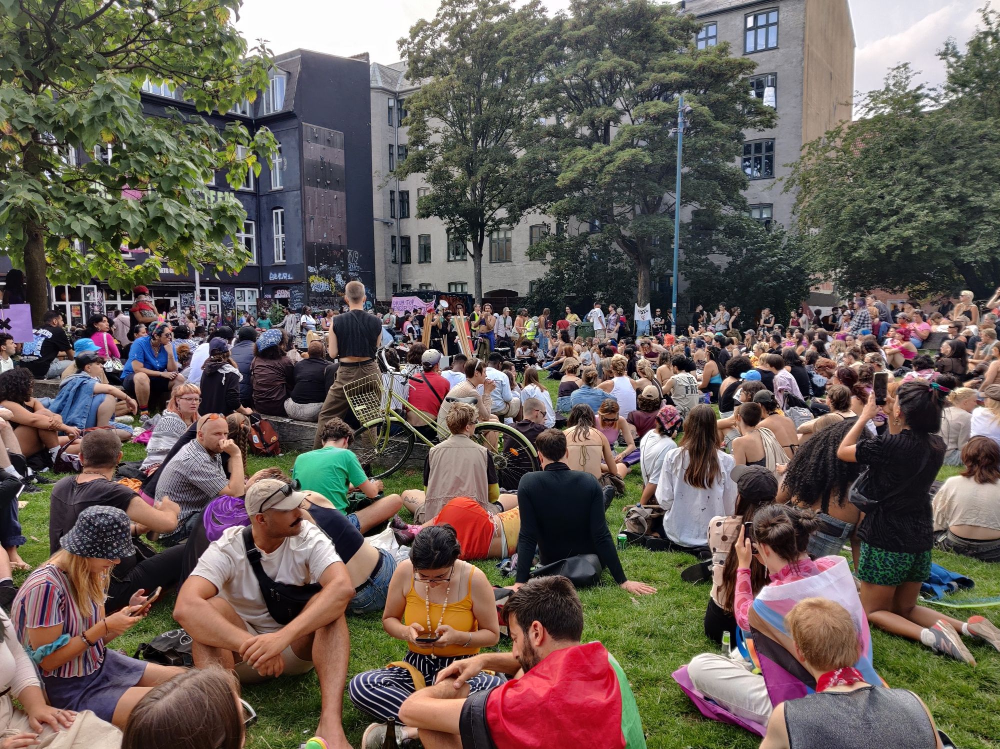 A crowd of queer people is sat in a park listening to appeals