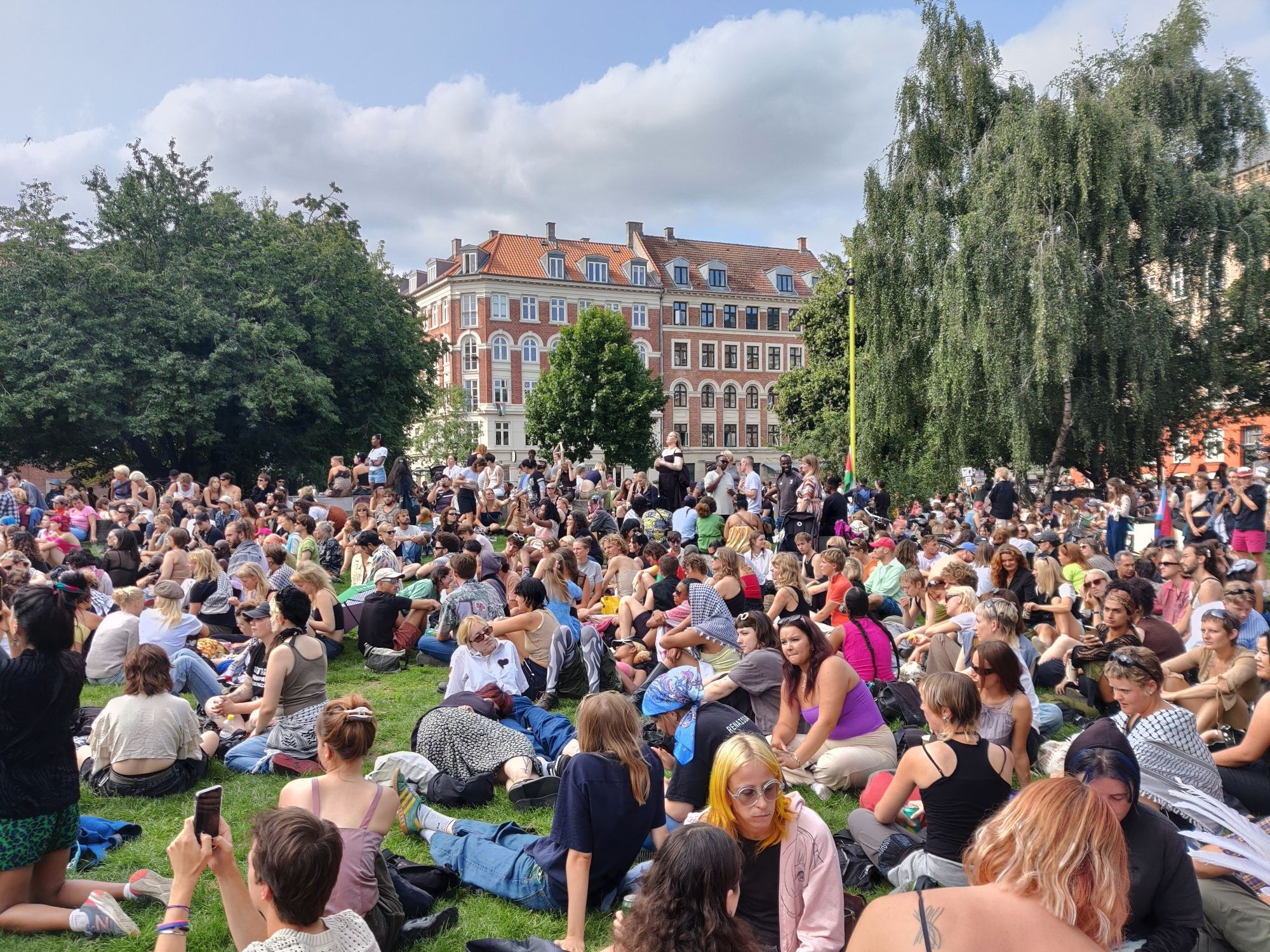 A crowd of queer people is sat in a park listening to appeals