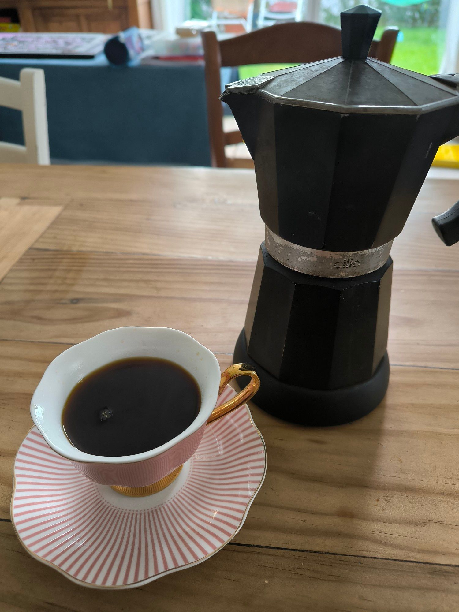 Sur une table en bois, une tasse de café en porcelaine et sa soucoupe, à côté d'une cafetière italienne noire. La tasse est vieux rose avec des accents dorés.